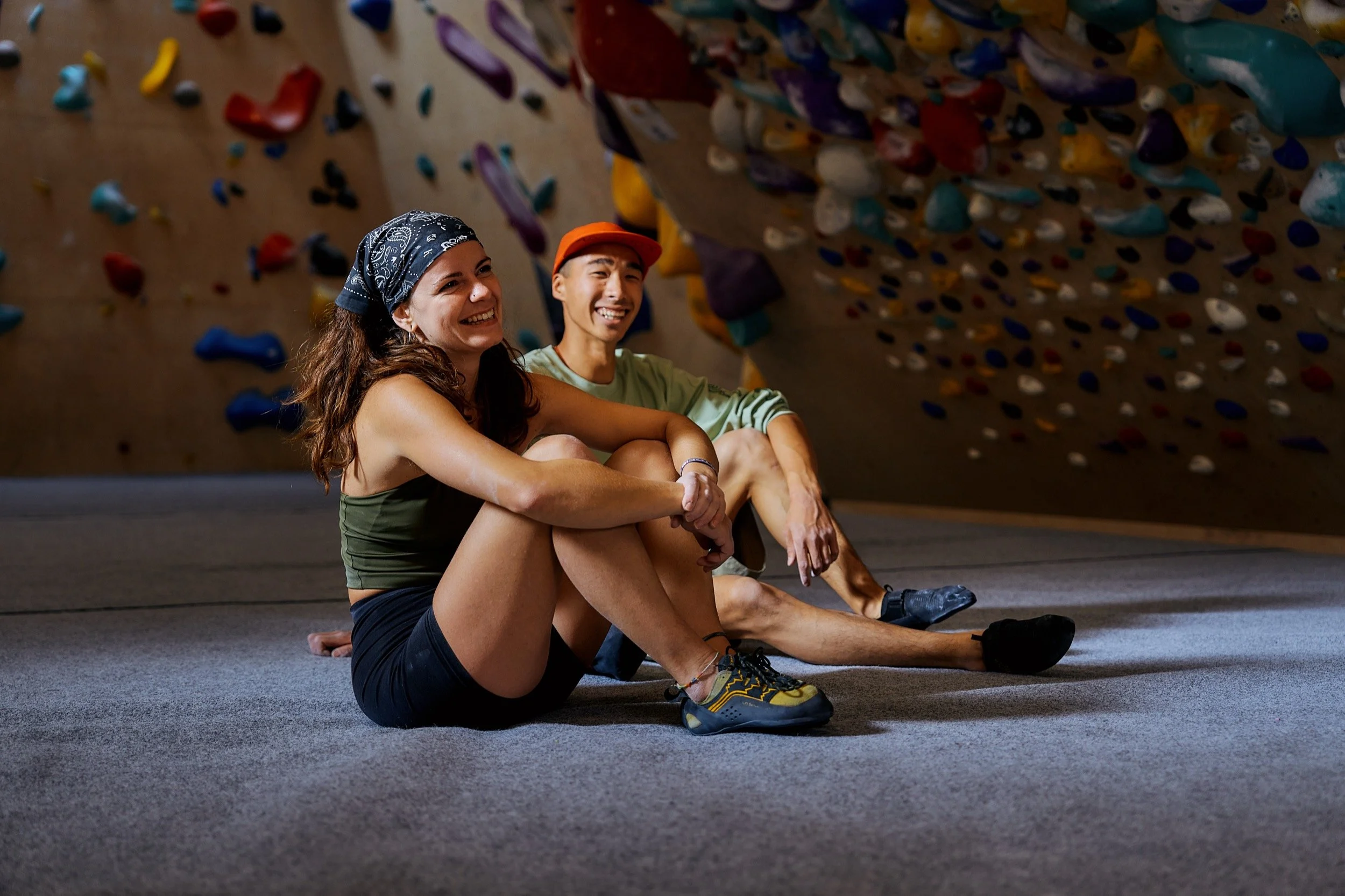 Zwei junge Menschen mit Kletterausrüstung sitzen lachend auf dem Boden eines Kletterhauses vor einer Wand mit bunten Griffen.