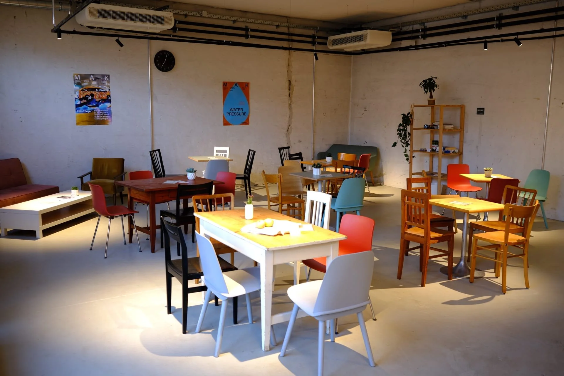 Cozy cafe interior with various colorful tables and chairs, some adorned with small potted plants, against white brick walls decorated with posters and a clock.