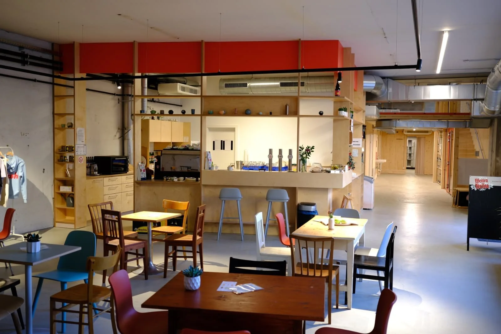 Interior of a modern cafe with colorful chairs and tables, a wooden counter area behind high stools, and open loft-style design.