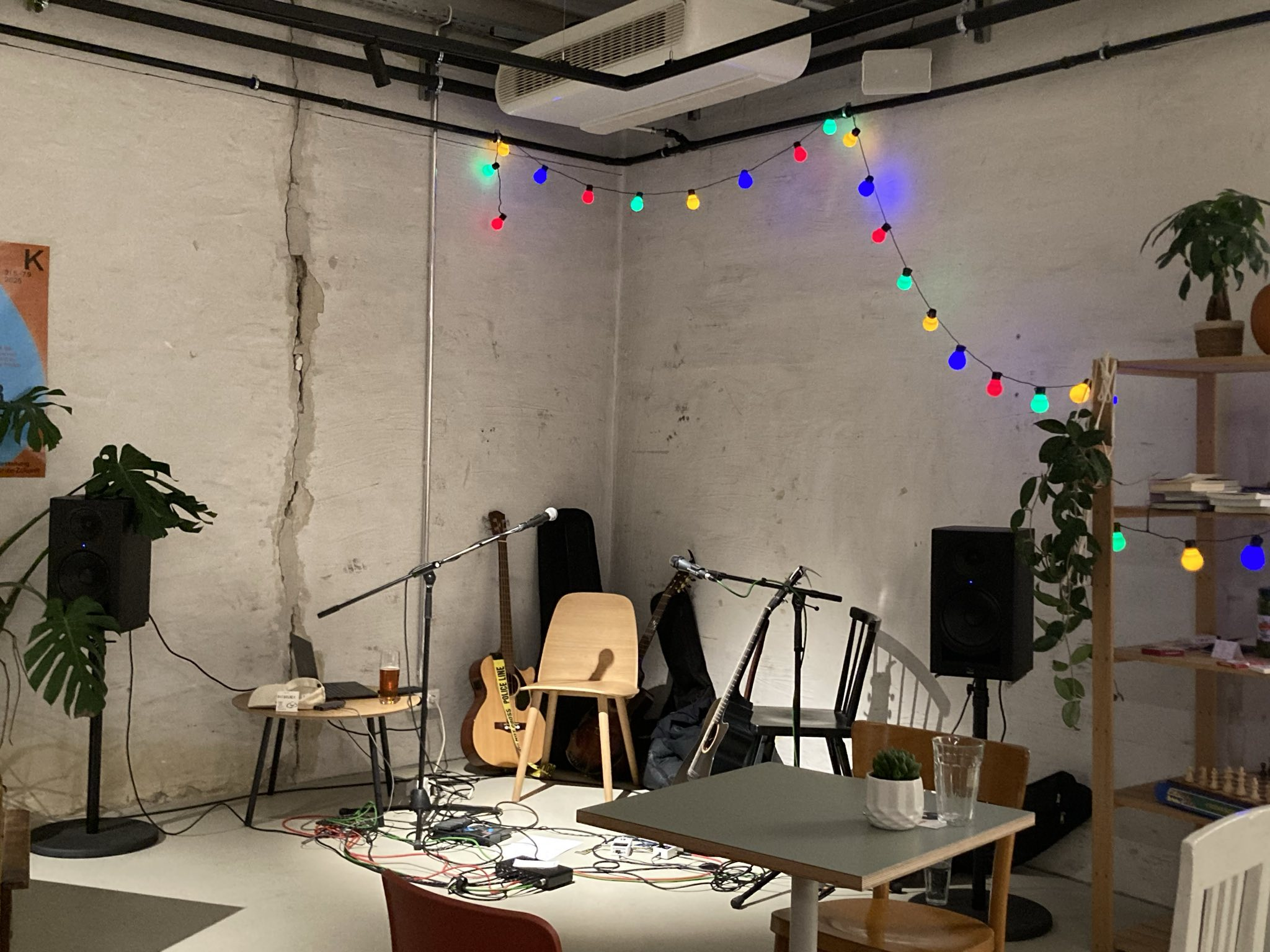 Empty music performance area with a microphone, guitars, speakers, and chairs, decorated with colorful string lights in a cozy, modern space.