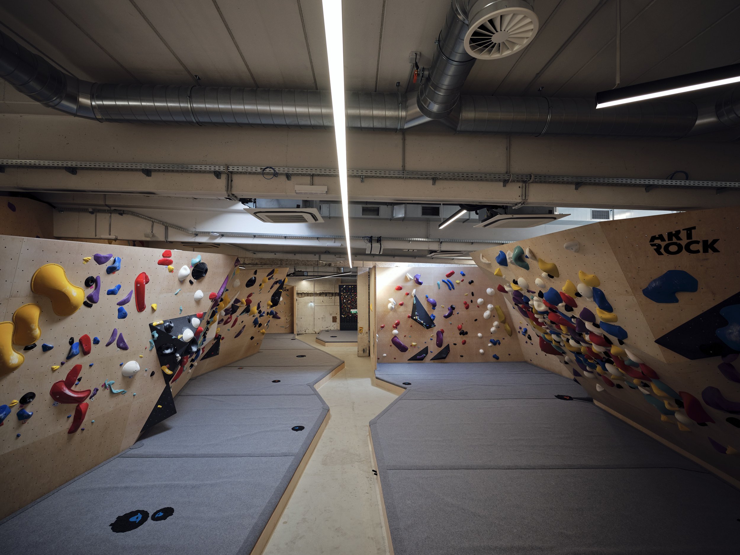 Innenansicht einer Kletterhalle mit bunten Klettergriffen an der Wand und weichen Matten auf dem Boden.