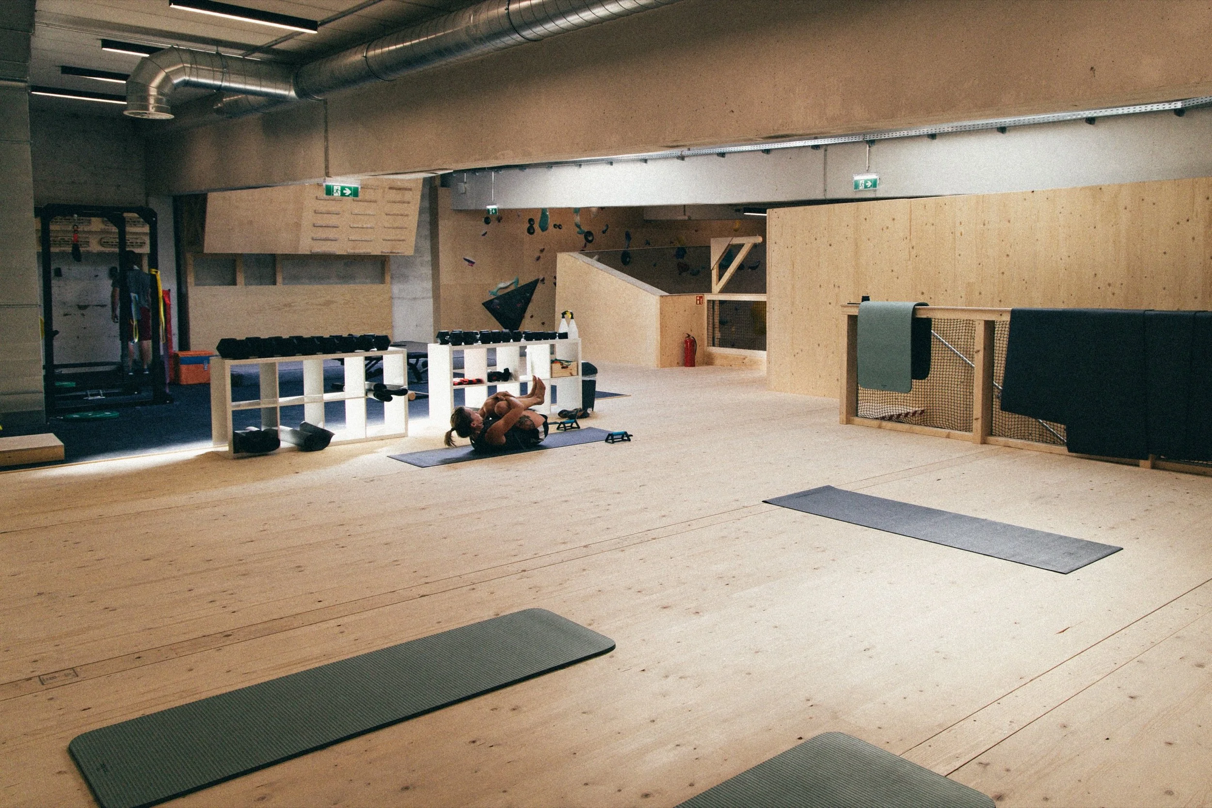 Ein leerer Indoor-Kletterraum mit Holzboden, Kletterwänden im Hintergrund, Trainingsgeräte und Yogamatten, sowie eine Frau, die im Vordergrund Yoga macht.