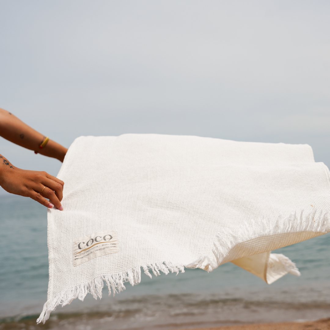 Persona que sosté una tovallola blanca amb la marca "COCO" a la platja, amb l'oceà i el cel ennuvolat al fons.