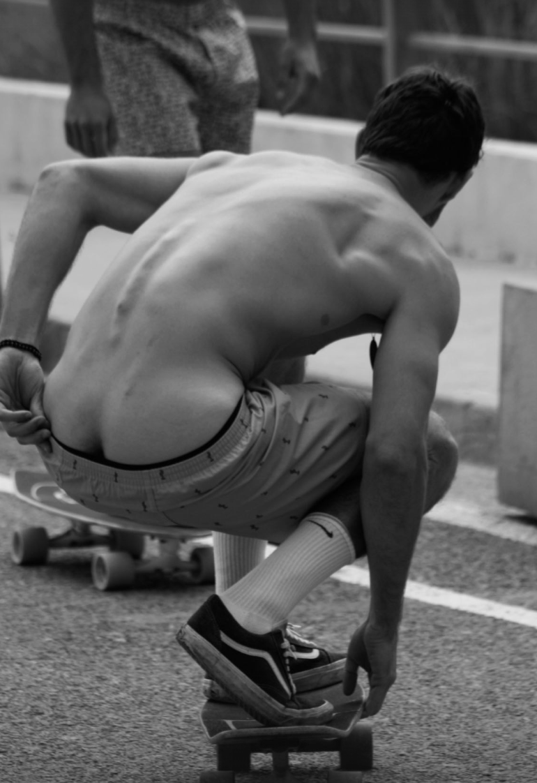 Un jove sense camisa amb els cabells curts i foscos ajupit sobre un monopatí, ajustant-se la sabata, en un parc de patinatge a l'aire lliure, amb una altra persona parcialment visible al fons.