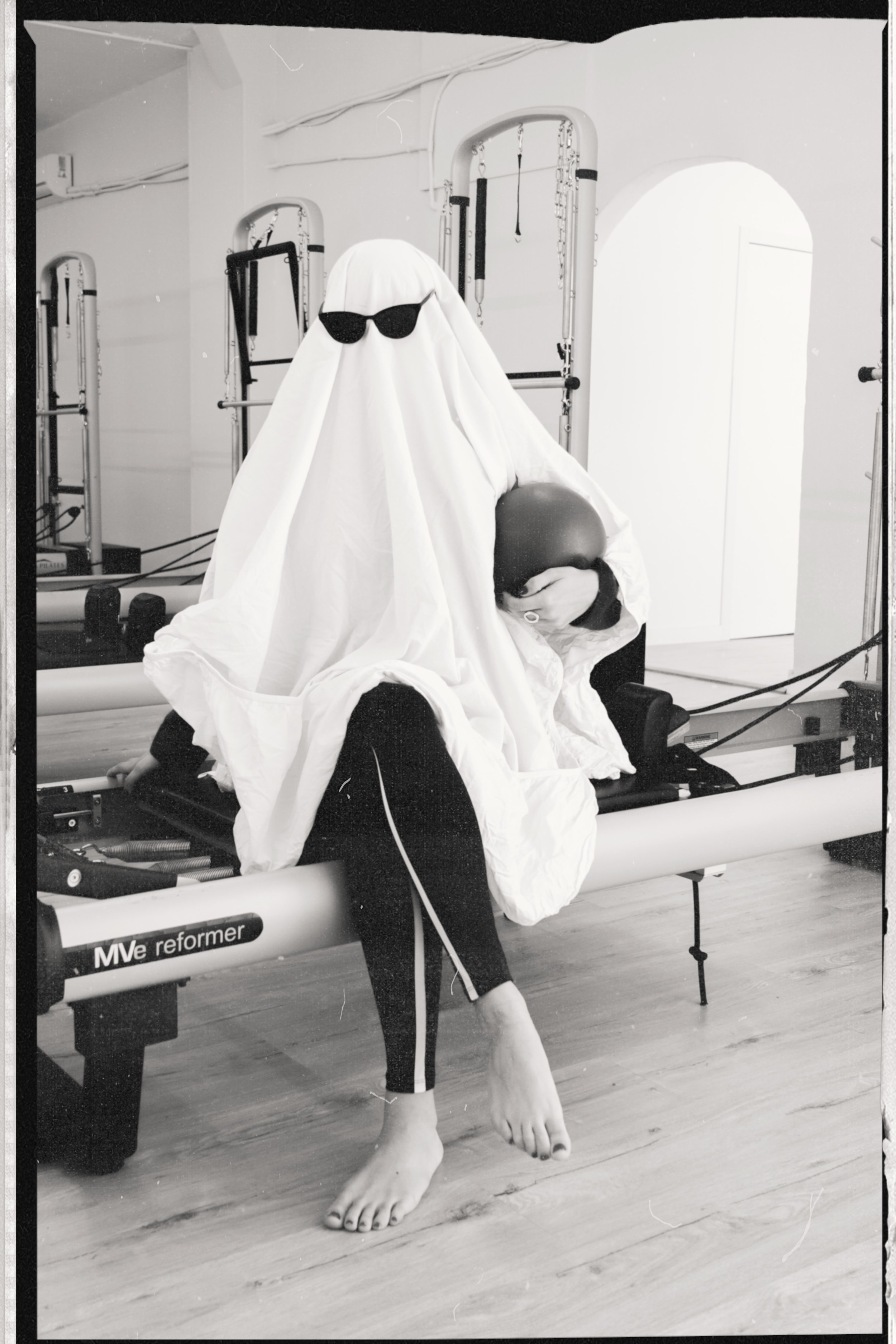 Una persona asseguda en una màquina de Pilates reformer, amb unes malles negres amb ratlles blanques, coberta per un llençol o manta blanca que s'assembla a una disfressa de fantasma, amb ulleres de sol afegides, en un gimnàs o estudi amb equipament 