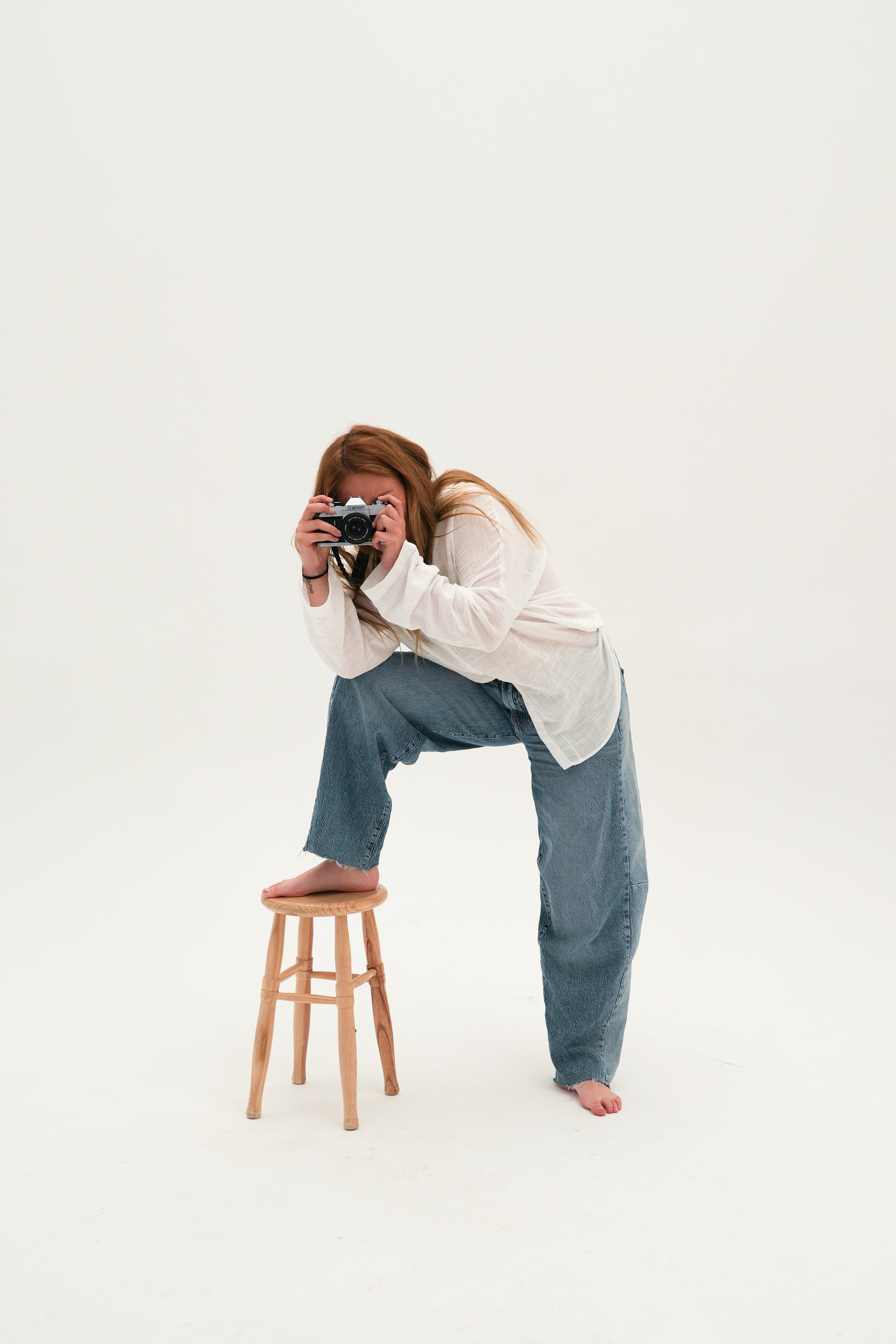 Persona amb els cabells llargs, amb camisa blanca i texans, ajupida amb un peu sobre un petit tamboret de fusta, sostenint una càmera davant de la cara, sobre un fons blanc llis.