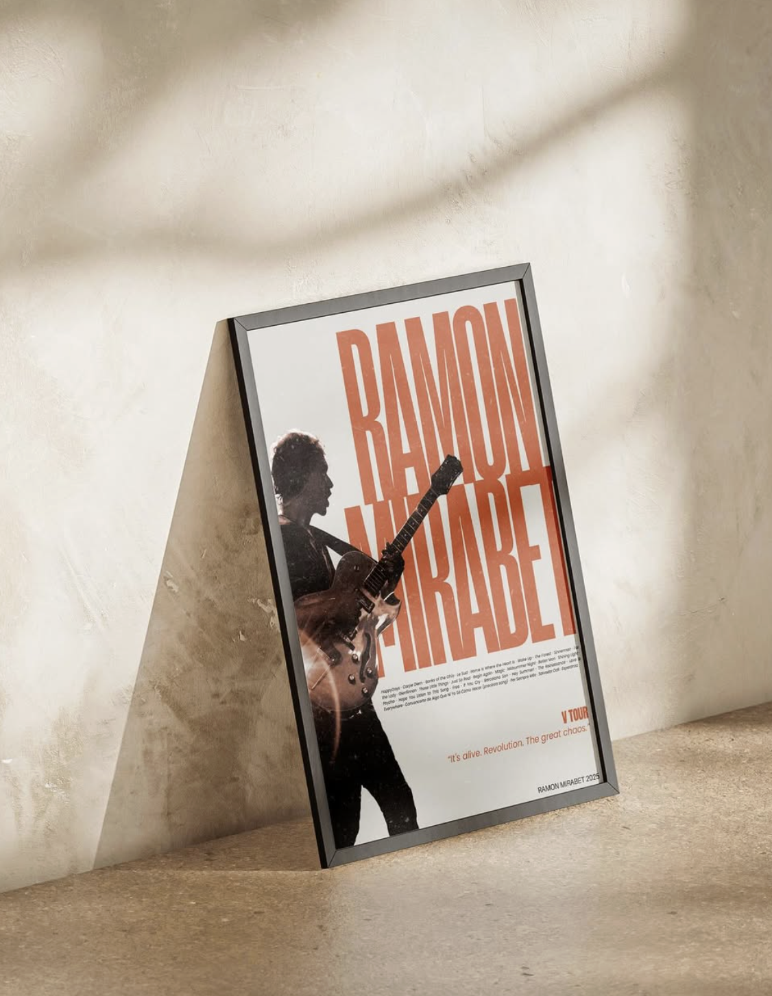 Pòster emmarcat del concert de RAMON MIRABET recolzat contra una paret i un terra beix, amb la silueta d'un guitarrista amb un text taronja gran i un text descriptiu més petit.