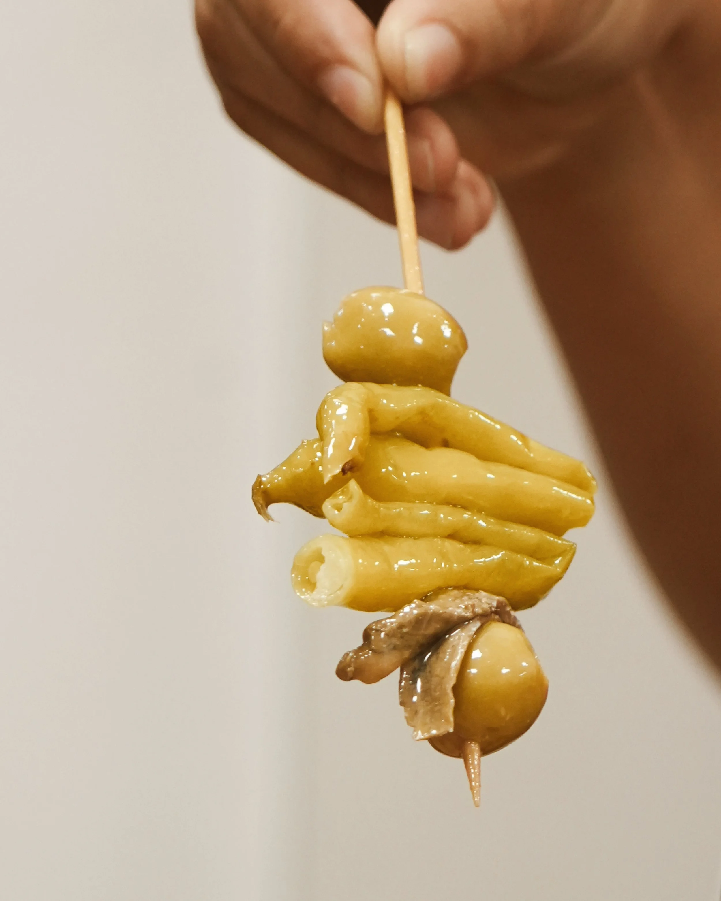 Persona que sosté una broqueta amb verdures en vinagre, com ara olives verdes, cors d'escarxofa i mongetes verdes, tot cobert amb una marinada brillant.