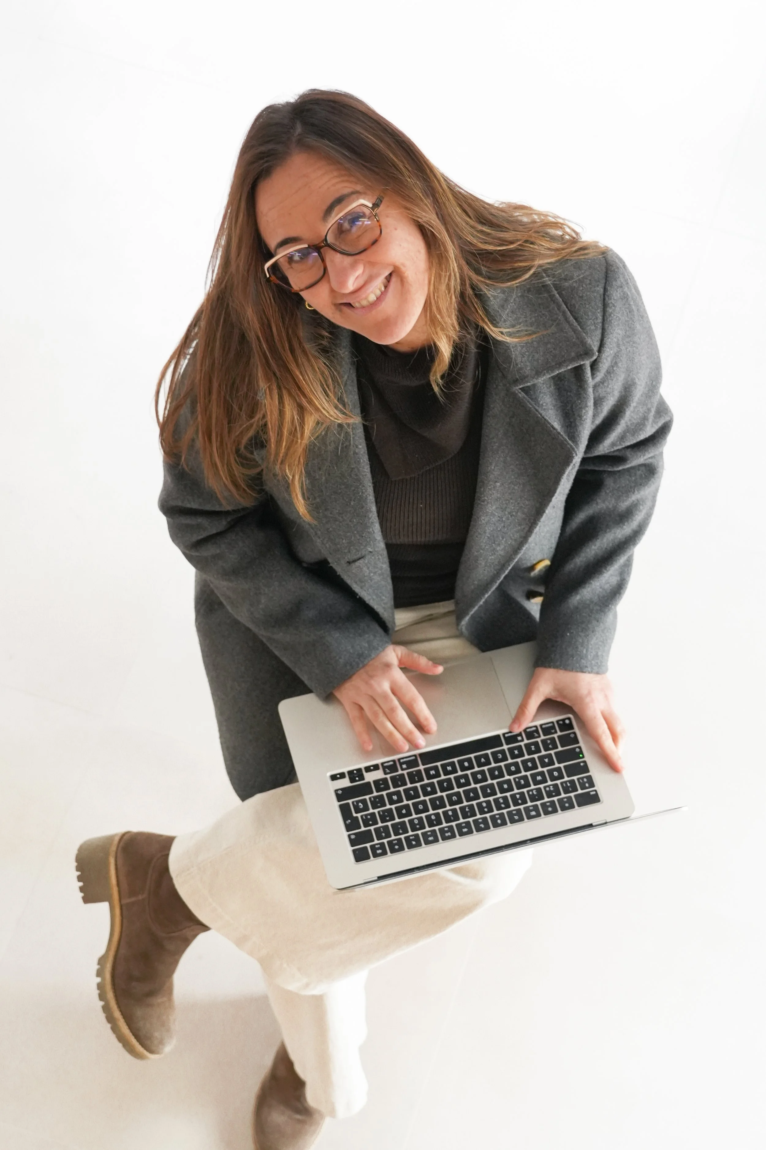 Una dona amb ulleres, cabells castanys, amb un abric gris fosc, una camisa negra, pantalons beix i botes marrons, asseguda en un tamboret blanc i treballant en un ordinador portàtil platejat.