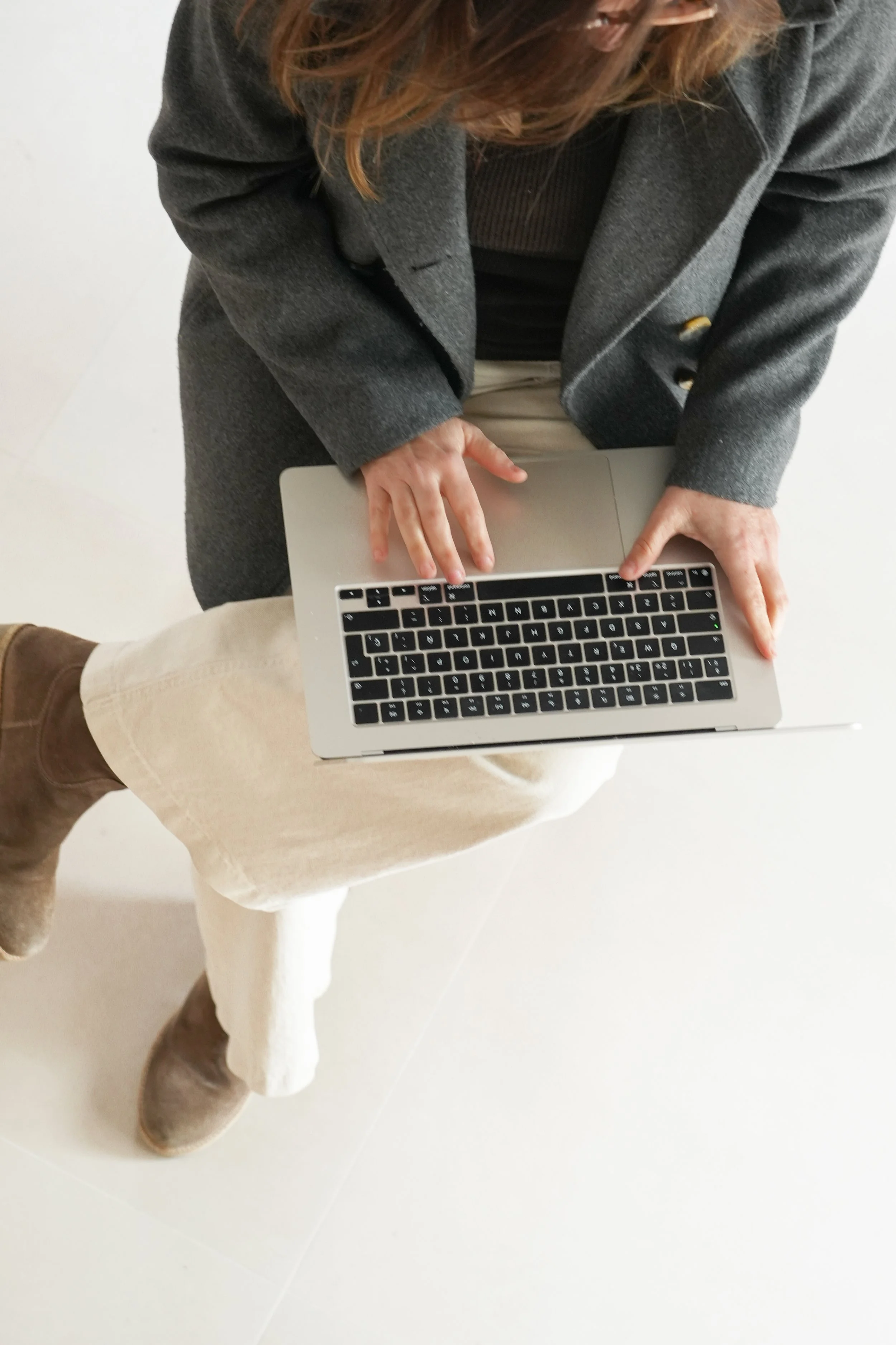 Una persona asseguda en una cadira blanca, treballant en un ordinador portàtil platejat, amb un abric gris fosc, pantalons beix i sabates marrons, vista des d'una perspectiva de dalt a baix.
