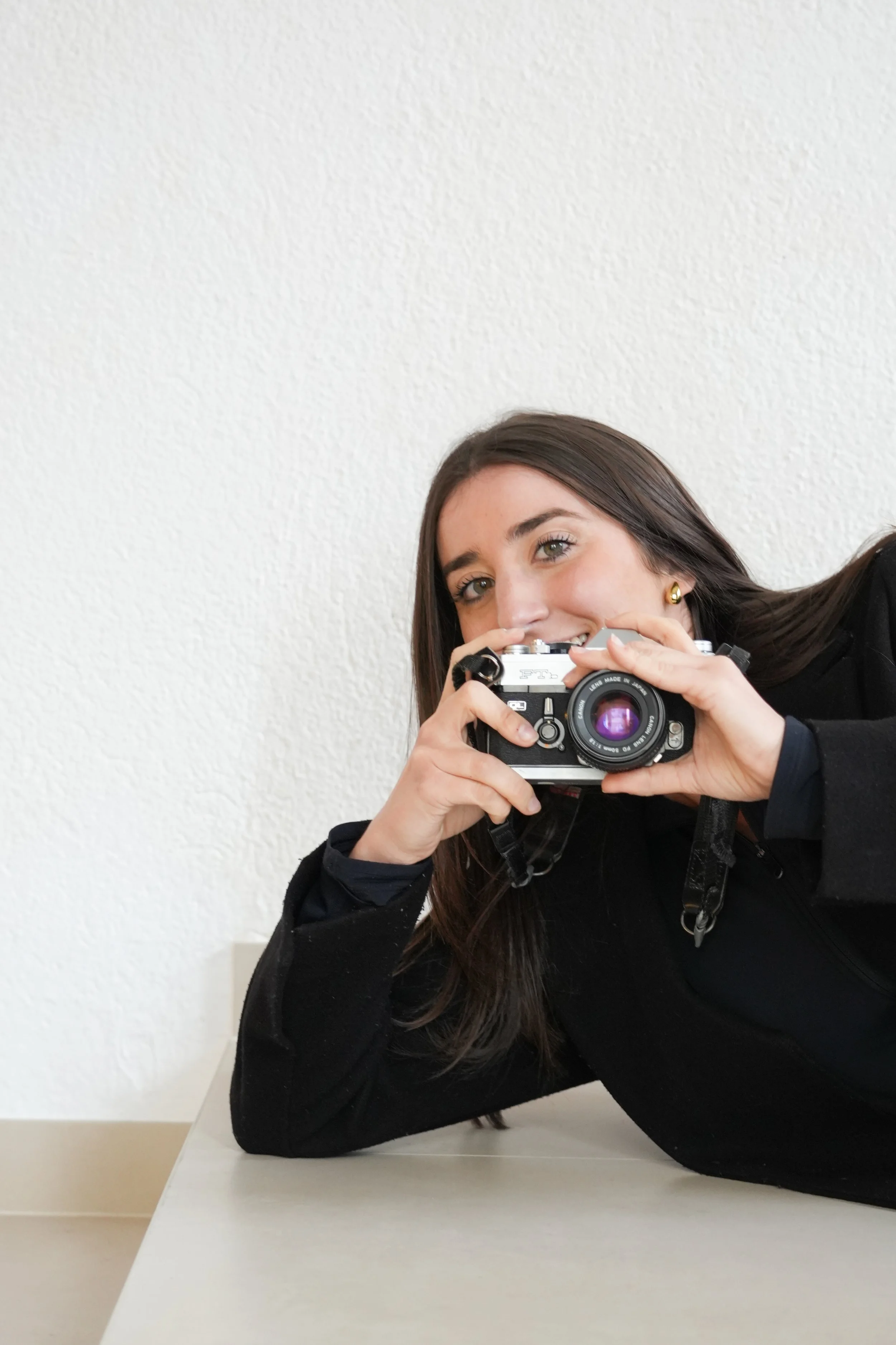Una dona jove amb els cabells llargs i foscos, amb una samarreta negra, sostenint una càmera davant de la cara i mirant cap a la càmera, recolzada a una taula en una habitació amb una paret blanca texturitzada.
