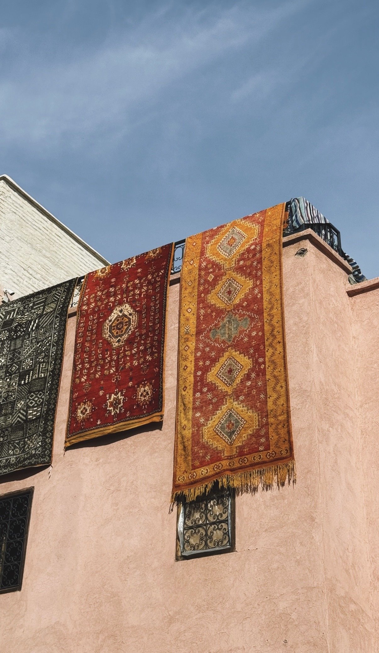 Catifes tradicionals de colors penjades sobre una paret d'edifici rosa amb petites finestres, sota un cel blau clar.