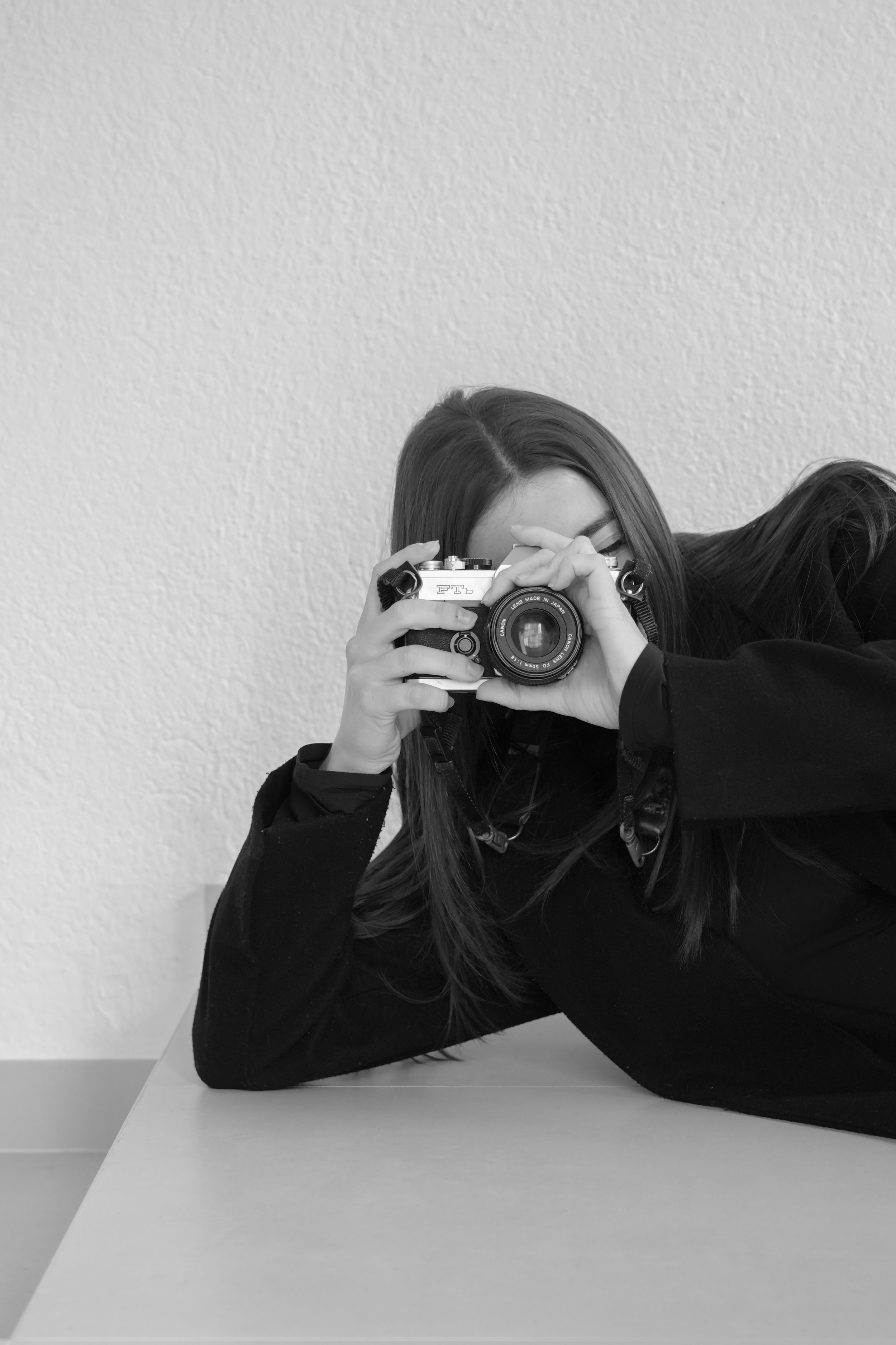 Una dona amb els cabells llargs i foscos estirada sobre una taula, fent una foto amb una càmera, contra una paret texturada.