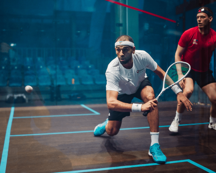 Two men playing squash on an indoor squash court, one is in a white shirt and black shorts, holding a squash racket near the ground, and the other is in a red shirt and black shorts, positioned behind.