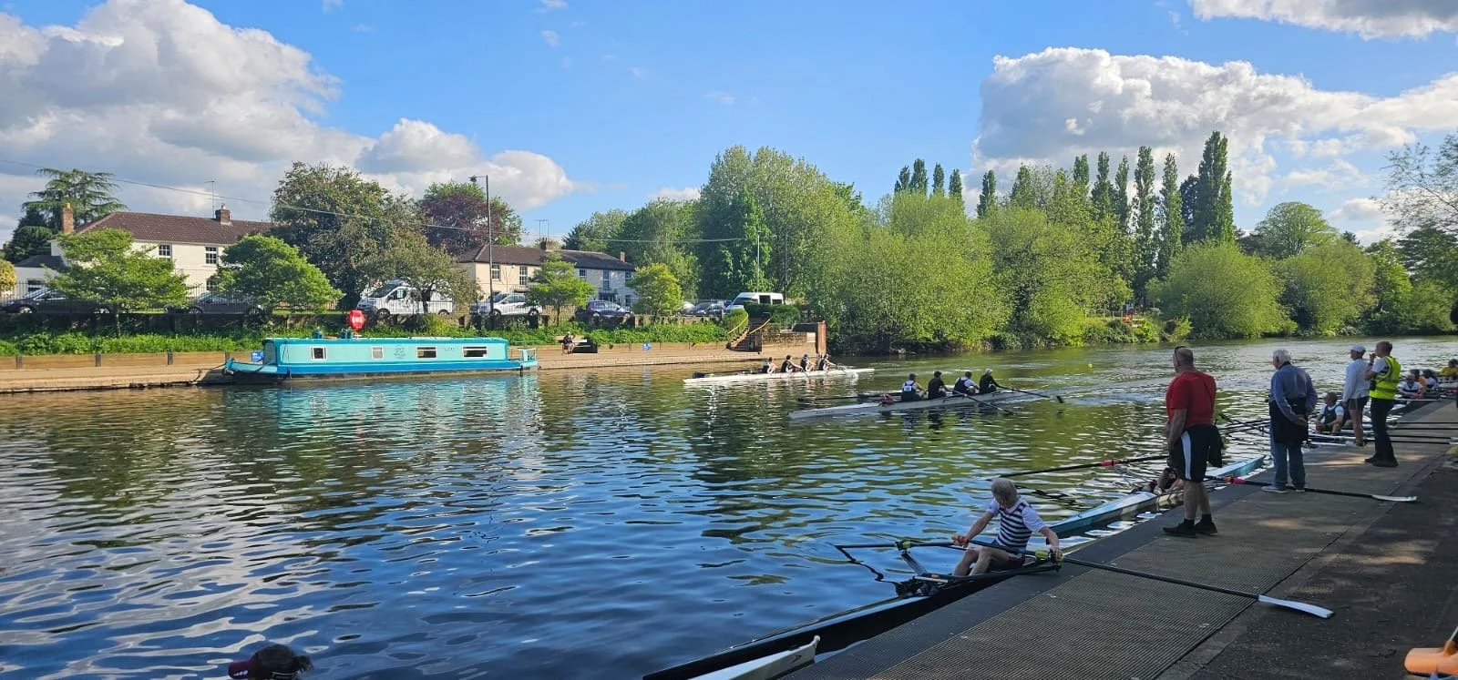 People rowing boats along a river on a sunny day with a blue sky and white clouds, trees lining the riverbanks, and houses in the background.