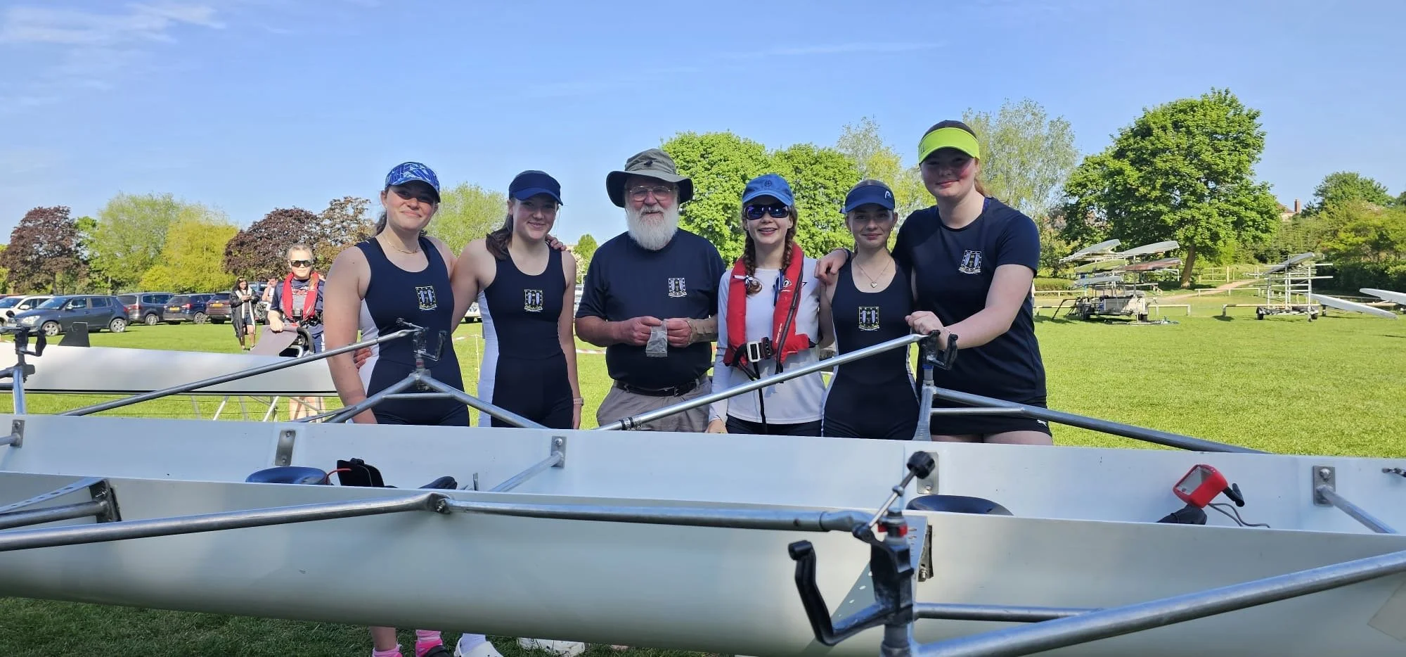 Group of six junior rowers and their coach, outdoors on a bright, sunny day at evesham regatta in 2025, with rowing boats and trees in the background.