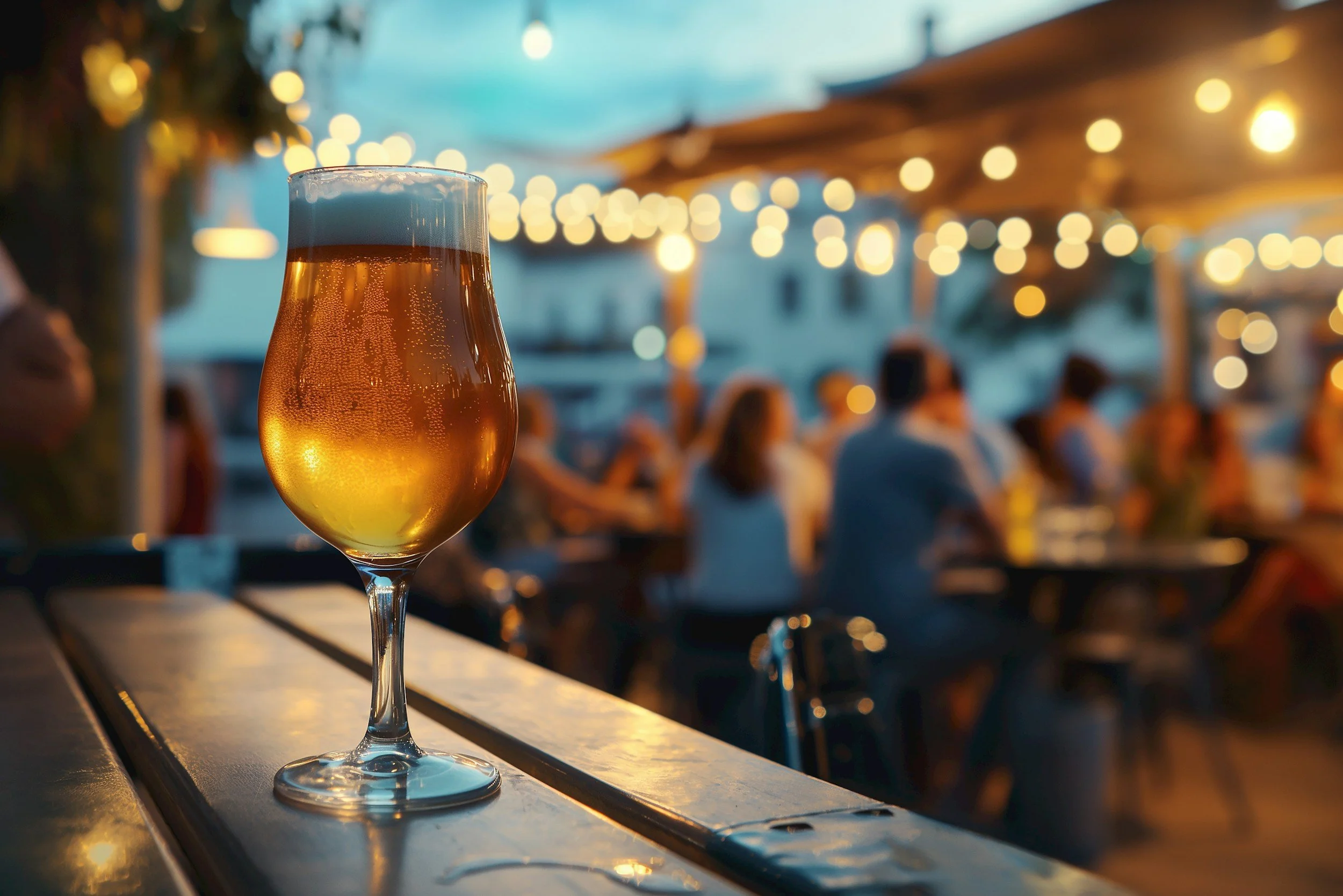 A glass of beer on a wooden table at an outdoor bar or restaurant during evening with string lights and people in the background.