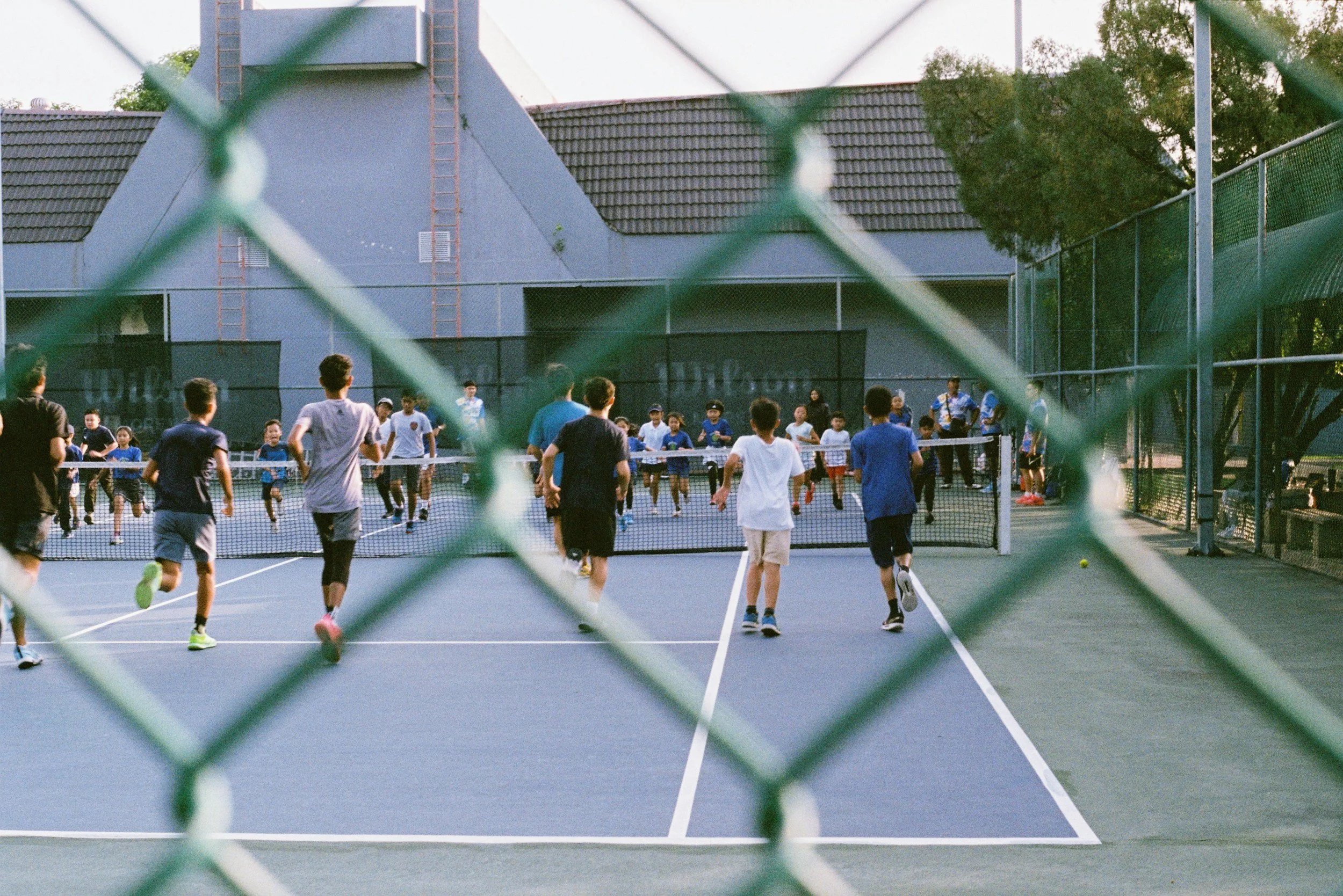Children and teenagers playing tennis on an outdoor court, viewed through a chain-link fence.