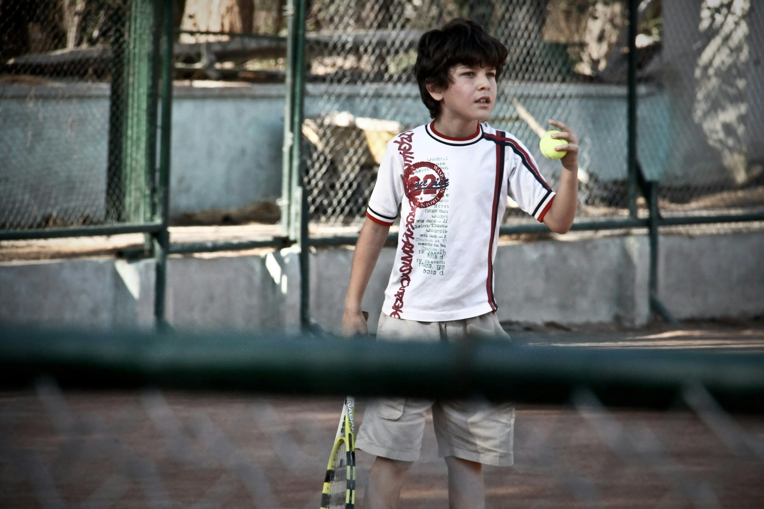 Young boy holding a tennis ball and a tennis racquet on a tennis court, surrounded by a chain-link fence.