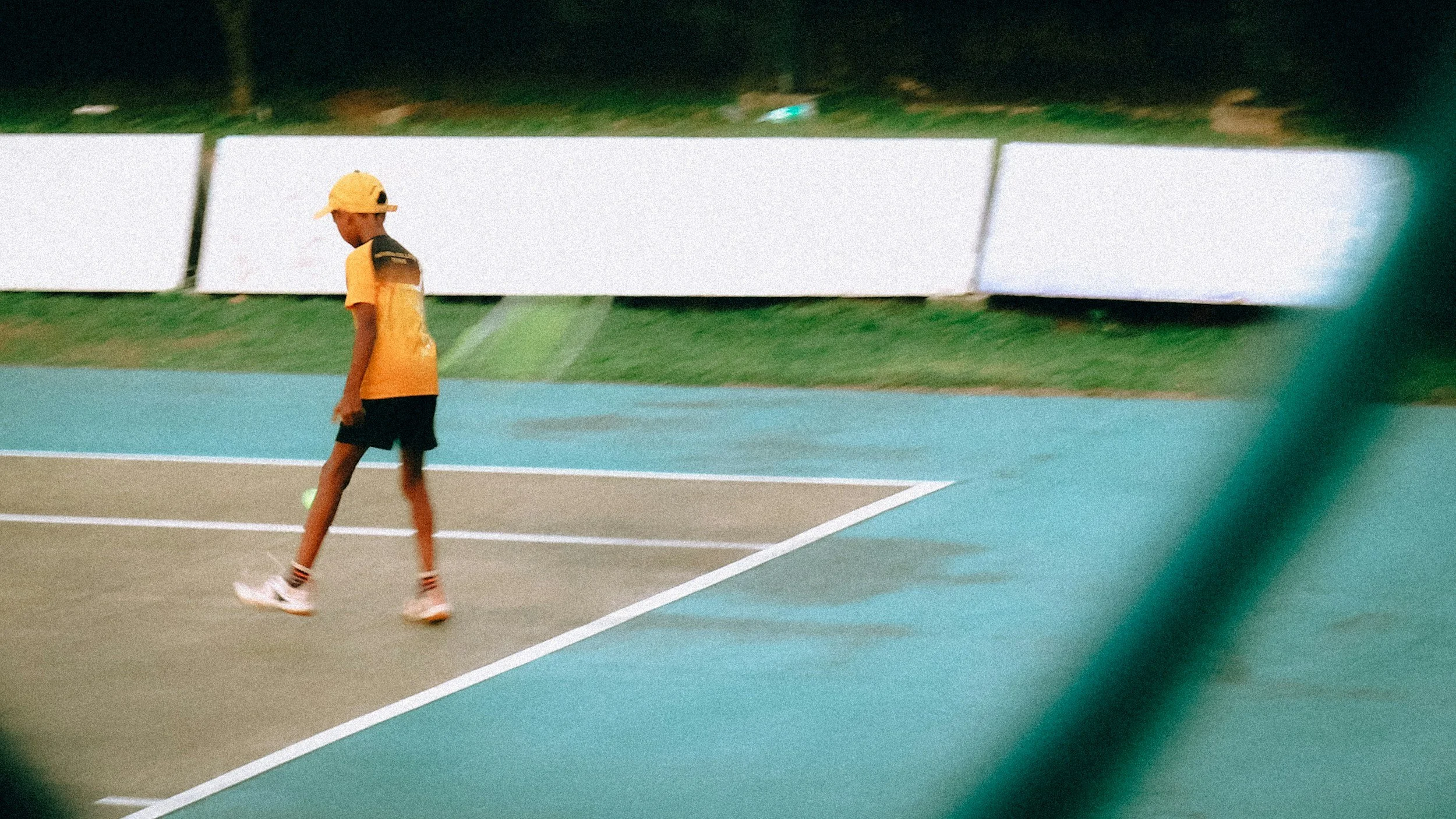 A young boy walking on a tennis court during daytime, wearing a yellow cap, yellow shirt, black shorts, and striped socks.