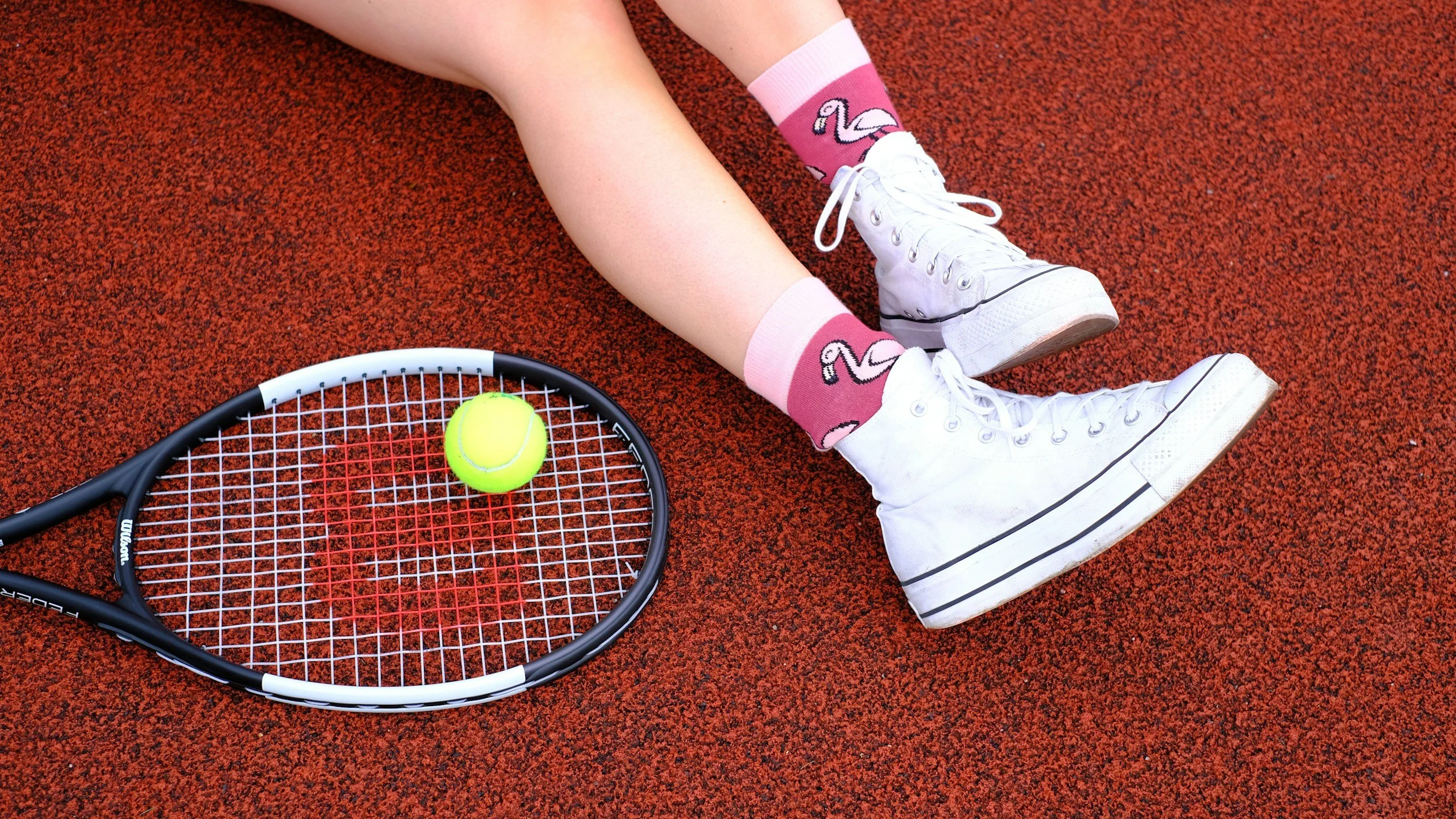 A child's legs are shown lying on a red tennis court, wearing white high-top sneakers and pink socks with flamingo designs. A tennis racket with a yellow tennis ball on top is placed beside them.