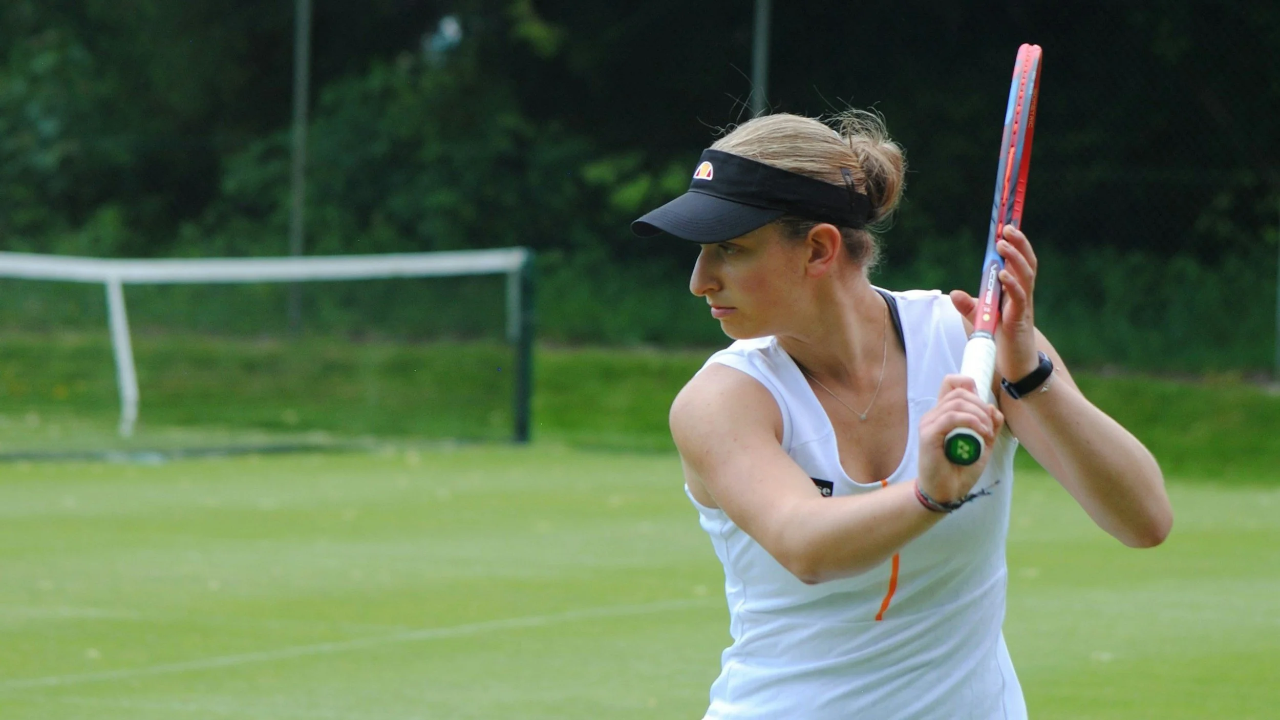 A woman practicing tennis on a grass court, holding a tennis racket, wearing a white sports top, black visor, and athletic accessories.