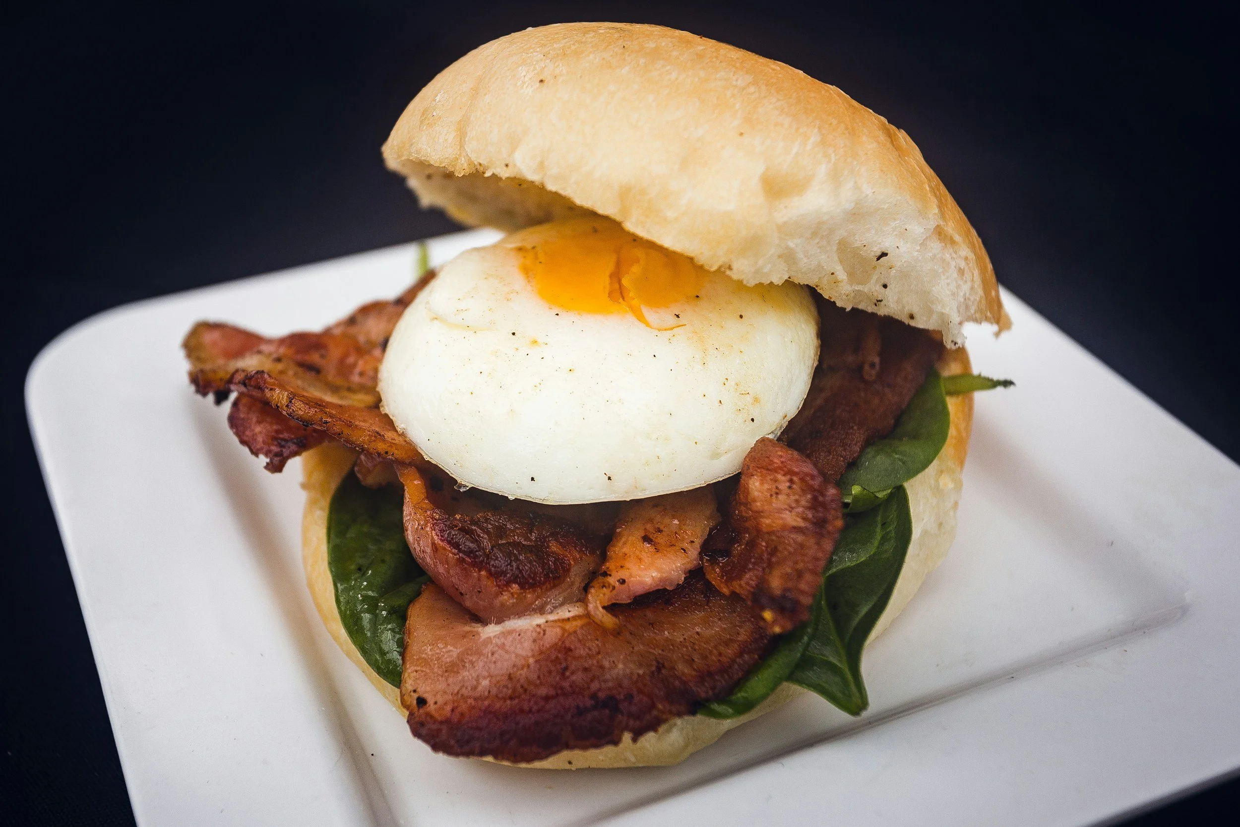 Bacon, greens, and a poached egg on a sandwich bun on a white plate