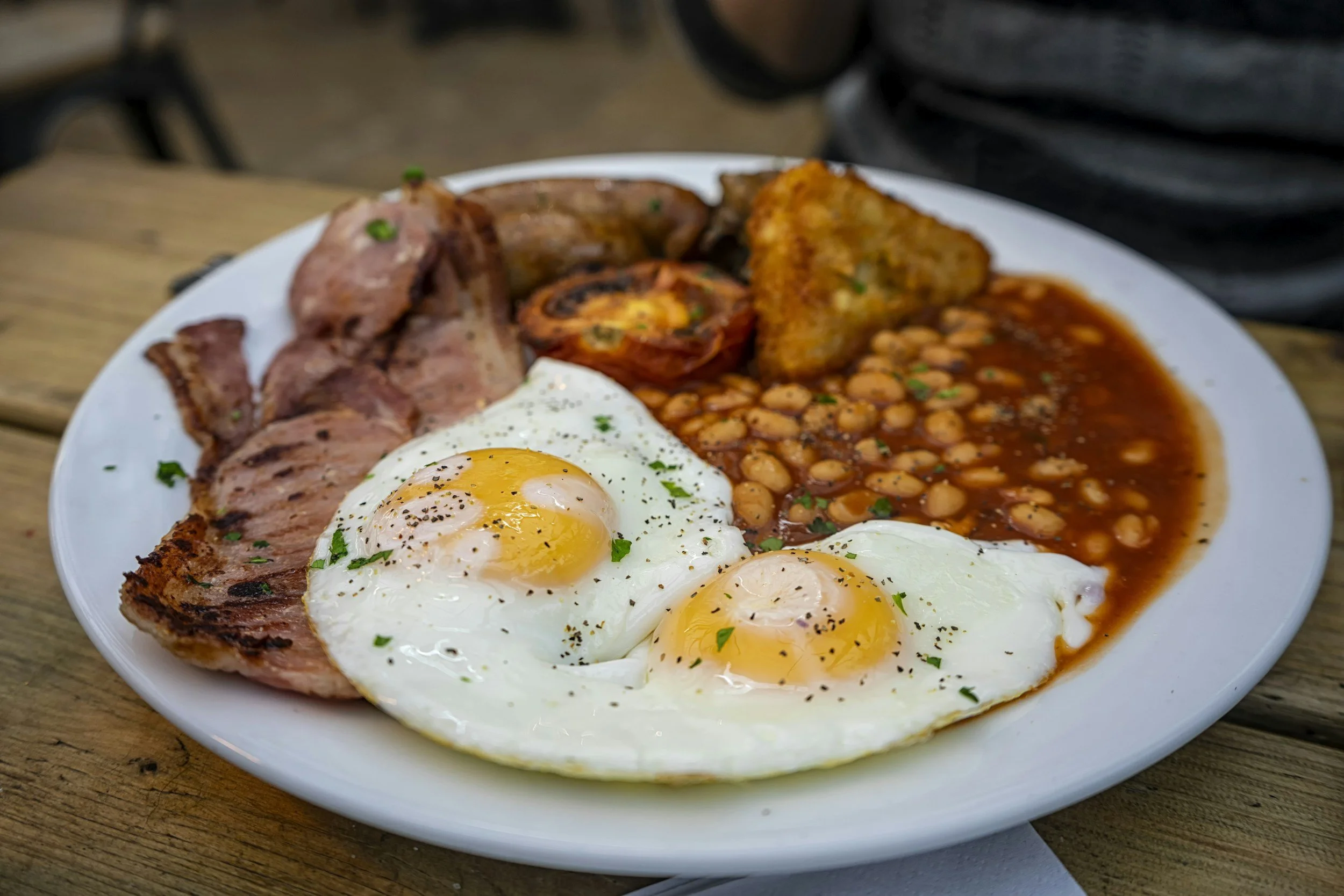 Plate of breakfast food including two sunny-side-up eggs, bacon, baked beans, fried tomato, hash brown, and sausage on a wooden table.