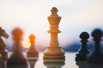 Close-up of a white chess king on a chessboard during sunset with blurred chess pieces in the background.
