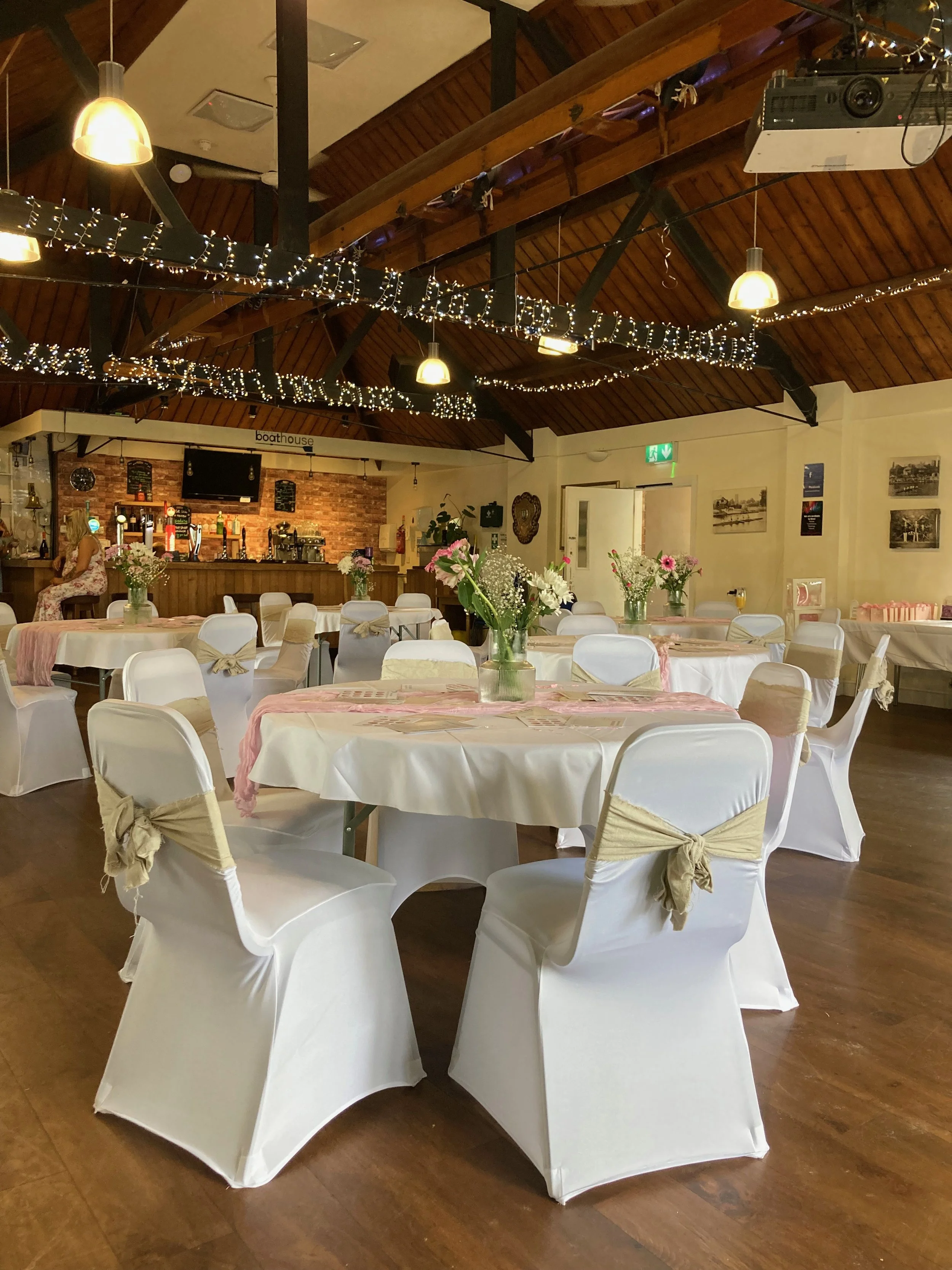 A decorated wedding venue at The Boathouse Cafe, part of Evesham Rowing and Racquets club, with round tables covered in white tablecloths, each with a floral center-piece, surrounded by white chairs with beige bows. 