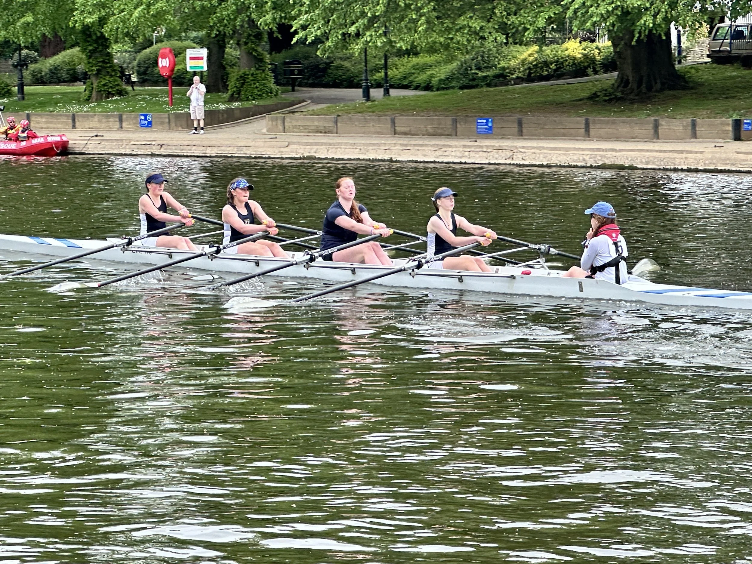 Four women in a rowing boat on the river avon, with a woman at the stern steering, and a person on shore in the background watching.