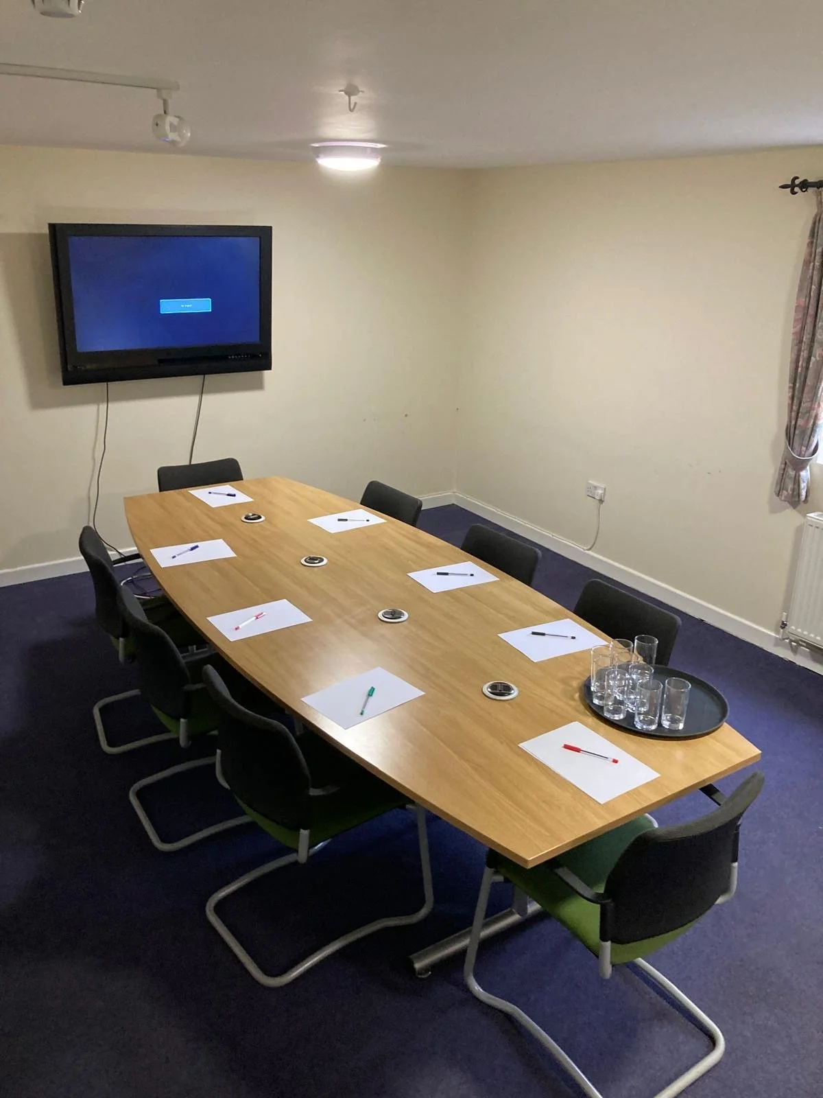 Meeting room with a rectangular table, eight chairs, a wall-mounted TV, and a tray with glasses on the table, with a pen and paper at each seat.