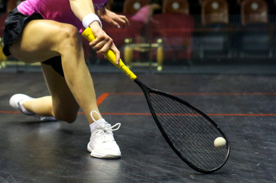 Person kneeling on a squash court, holding a squash racket and hitting a squash ball.