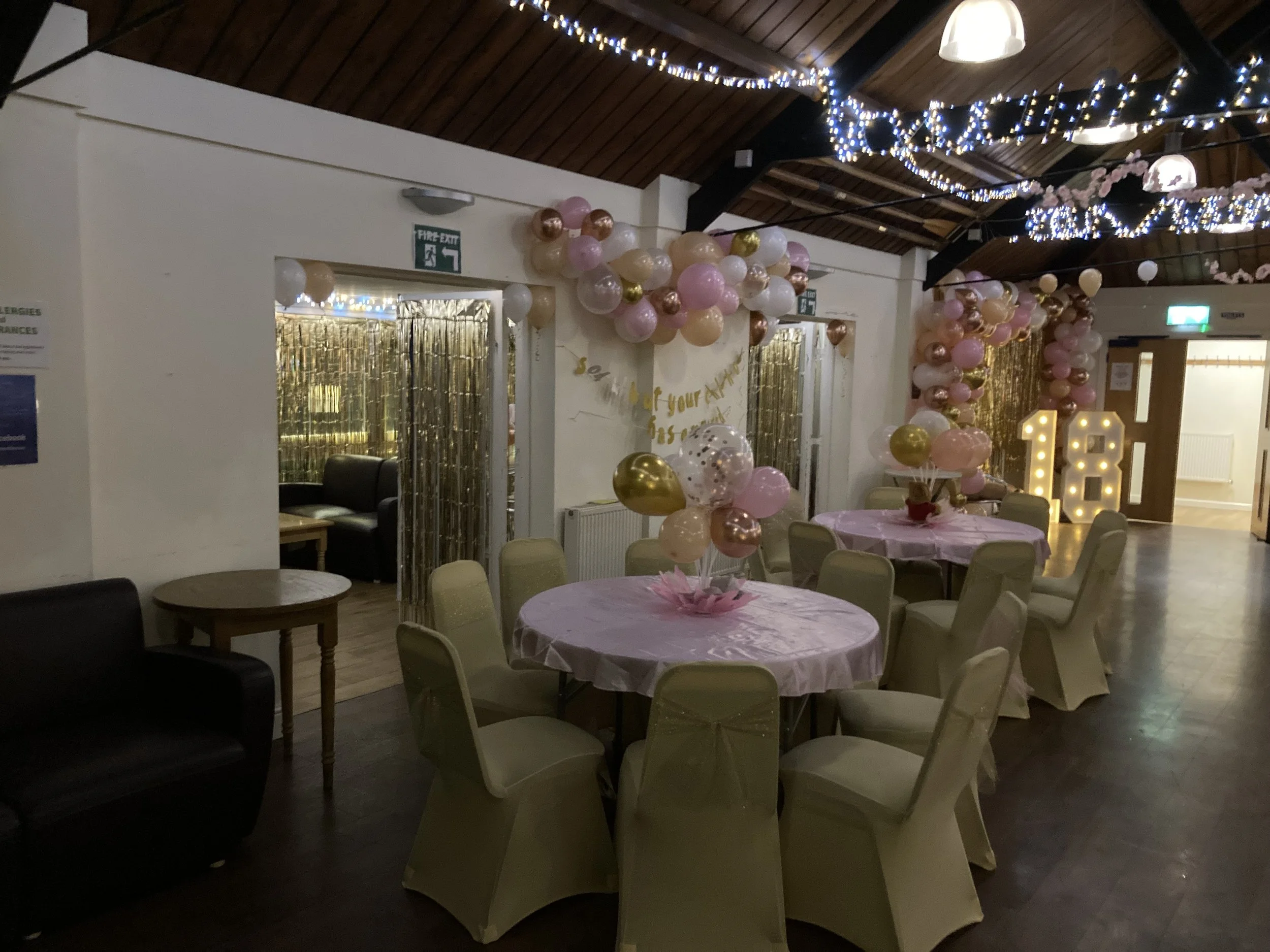Decorated event space with pink, white, gold, and transparent balloons, pink tablecloths, and a large light-up '18' sign, likely for an 18th birthday celebration.