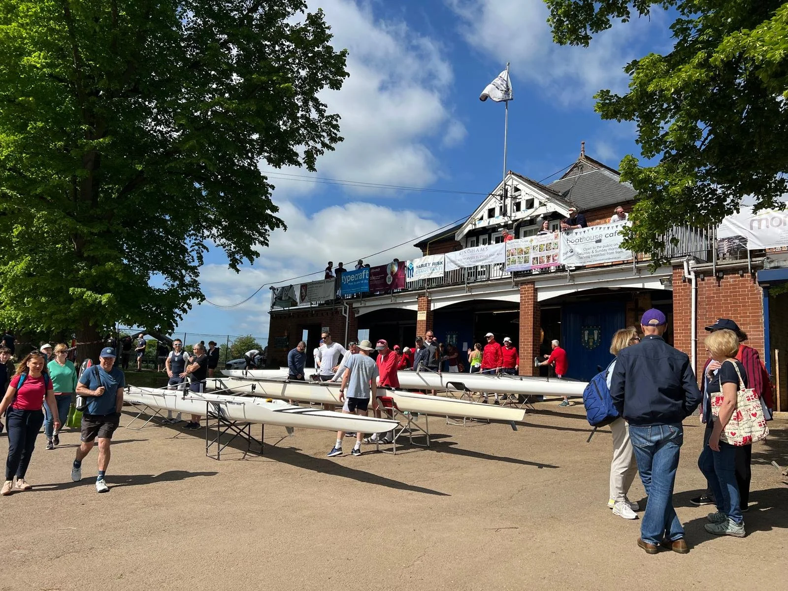 People gathered outside a boathouse with rowing shells on racks, some people sitting on the upper balcony, trees and blue sky with clouds in the background.