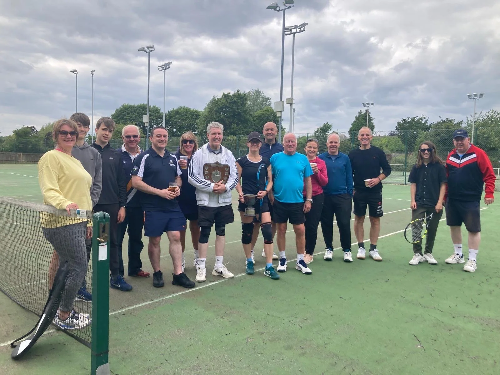 A group of people standing on a tennis court at Evesham Tennis Club, holding drinks and trophies, with cloudy sky above.