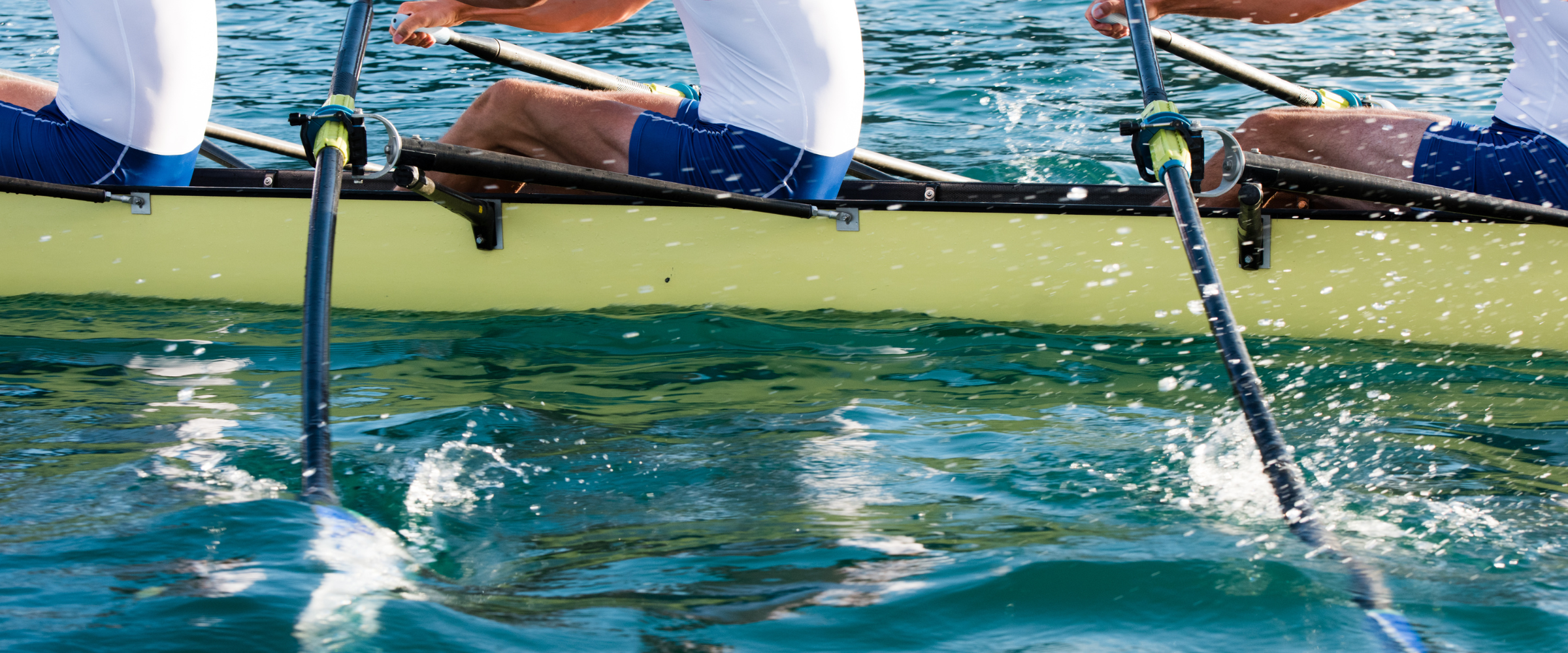 Partial view of a rowing team in a yellow boat, with team members wearing white shirts and blue shorts, rowing on calm water.