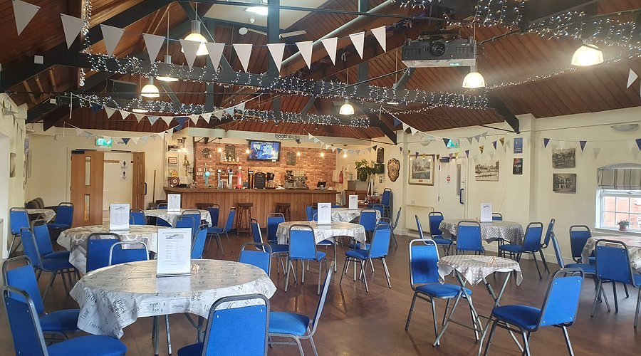 Empty event or dining hall with round tables covered in white tablecloths, along with blue chairs, a bar area at the back with a television, and decorations including bunting and string lights hanging from a wooden ceiling.