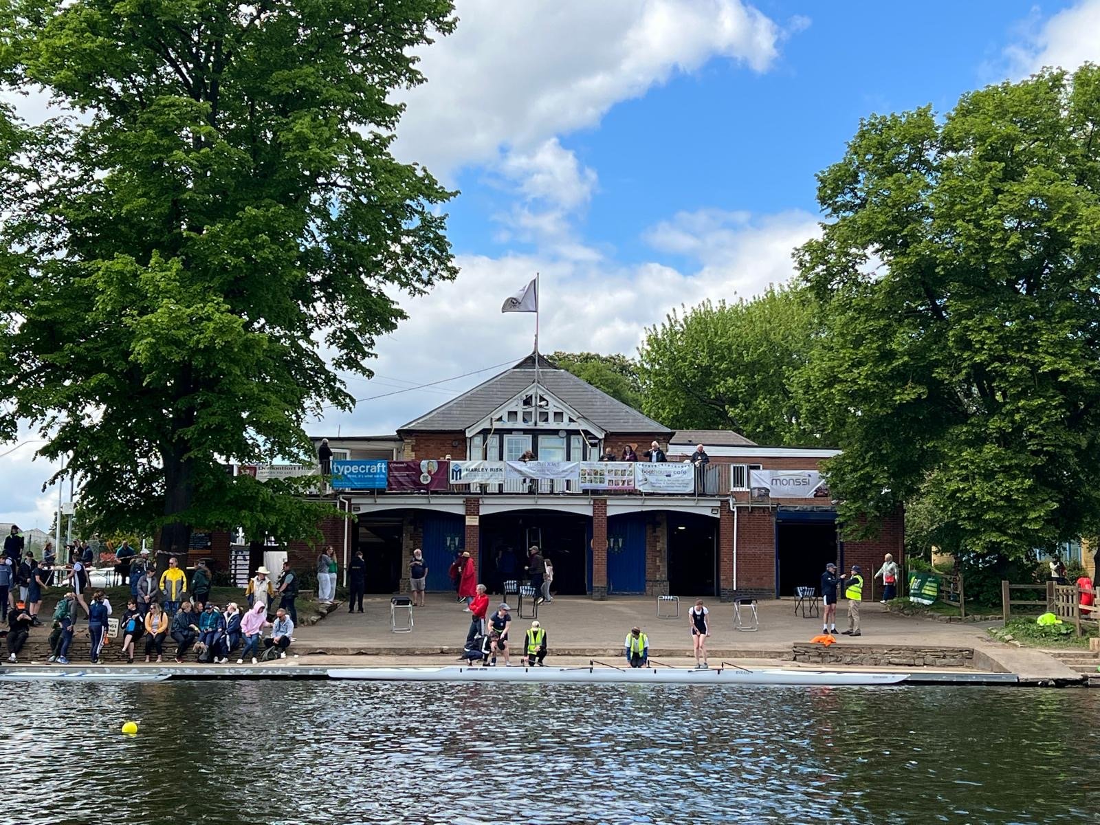 People gathered by the water at a riverside venue with a building featuring banners, trees, and a partly cloudy sky.