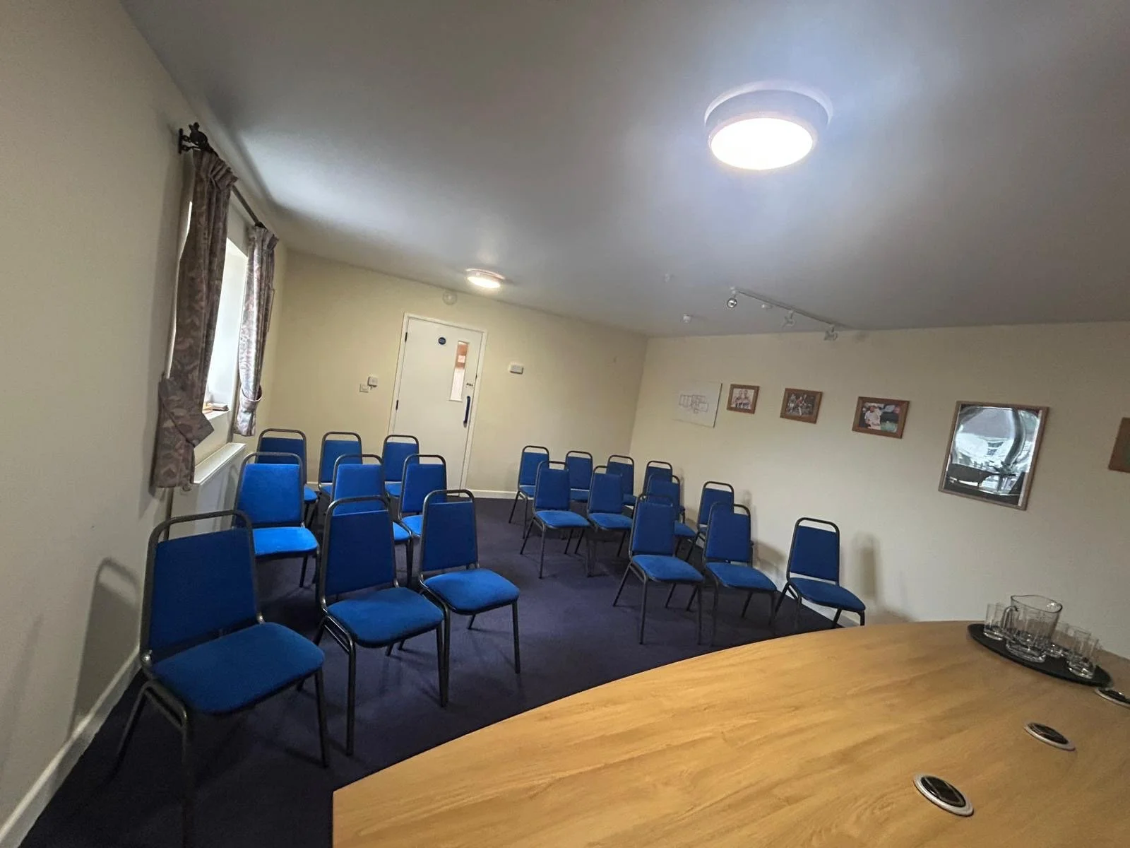 A small conference room with 14 blue chairs arranged in rows, windows with curtains on the left, a door at the back, framed pictures on the wall, a wooden table in the foreground with glasses on a tray, and ceiling lights.