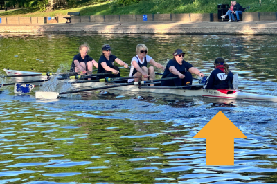 Five people rowing a boat on the river avon at evesham rowing and racquets club with trees and a riverside walkway in the background.
