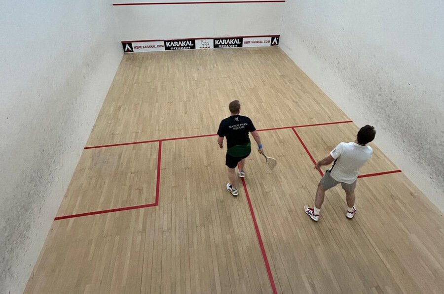 Two people playing squash on an indoor court atEvesham Squash Club with wooden flooring and red boundary lines.