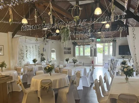 Event space decorated with white tablecloths, chairs with bows, floral centerpieces, string lights, and hanging greenery, set up for a celebration or reception.