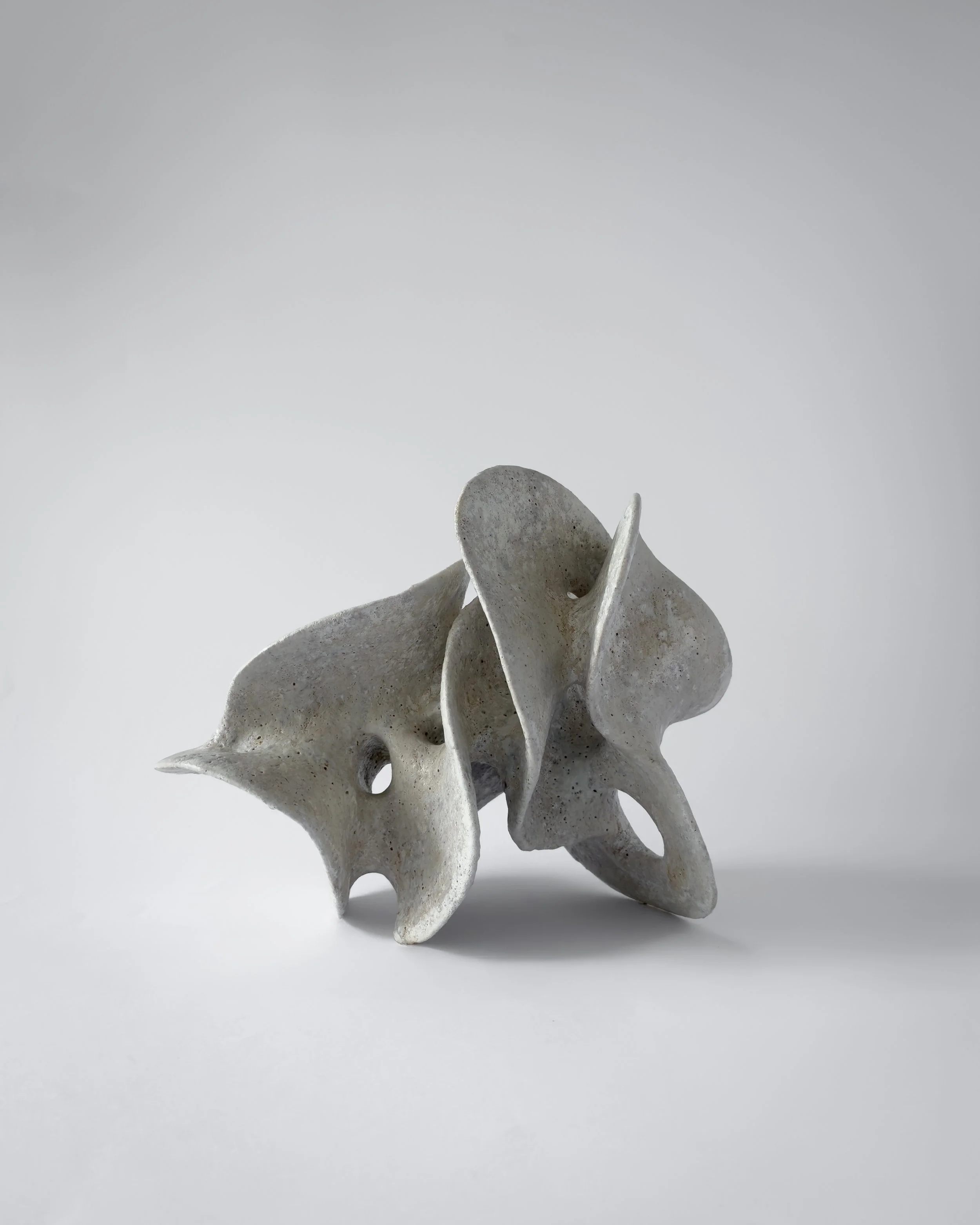 Abstract sculpture made of light-colored, porous material with organic, flowing shapes and holes, set against a plain white background.