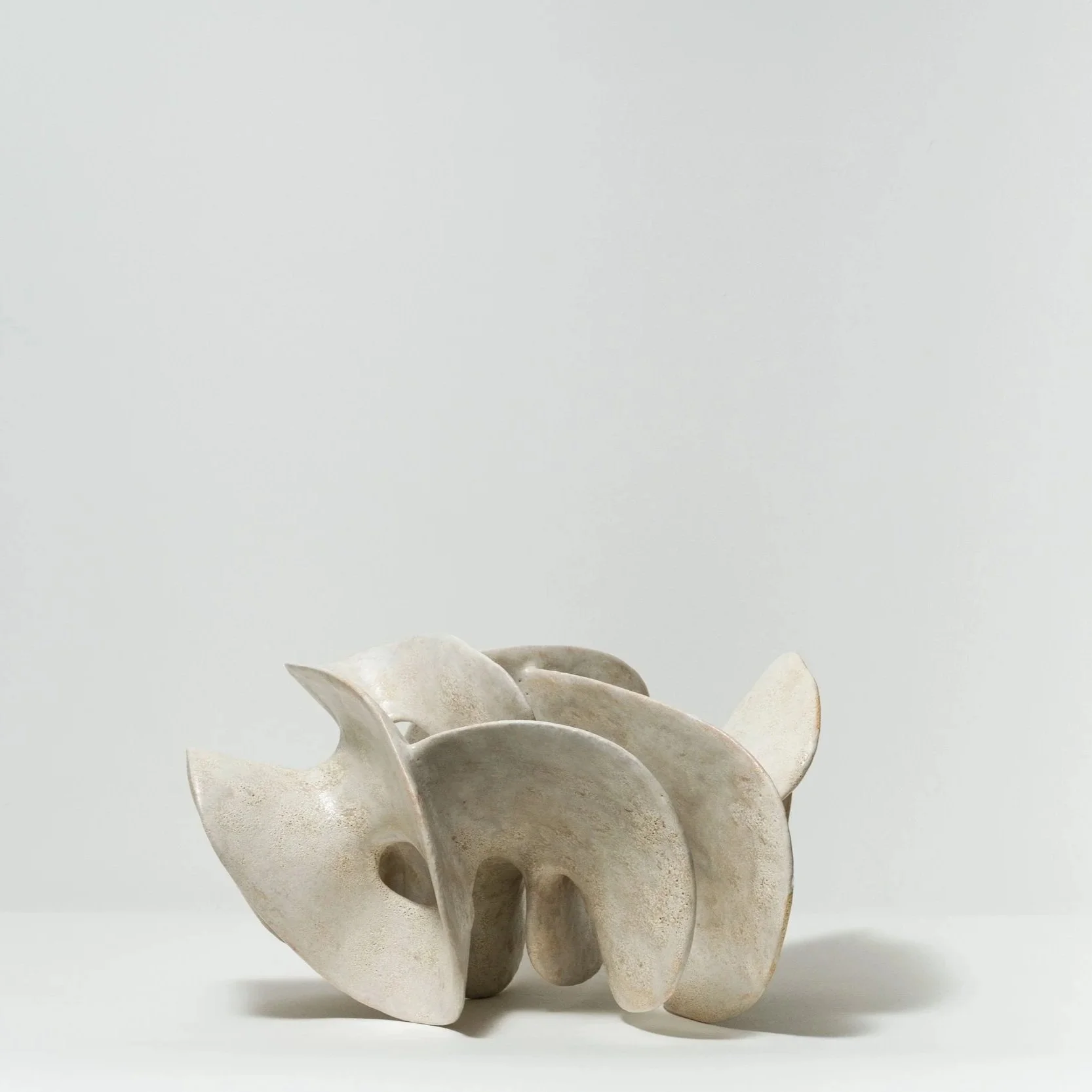 Abstract ceramic sculpture with curved, organic shapes in a light beige color on a white background.