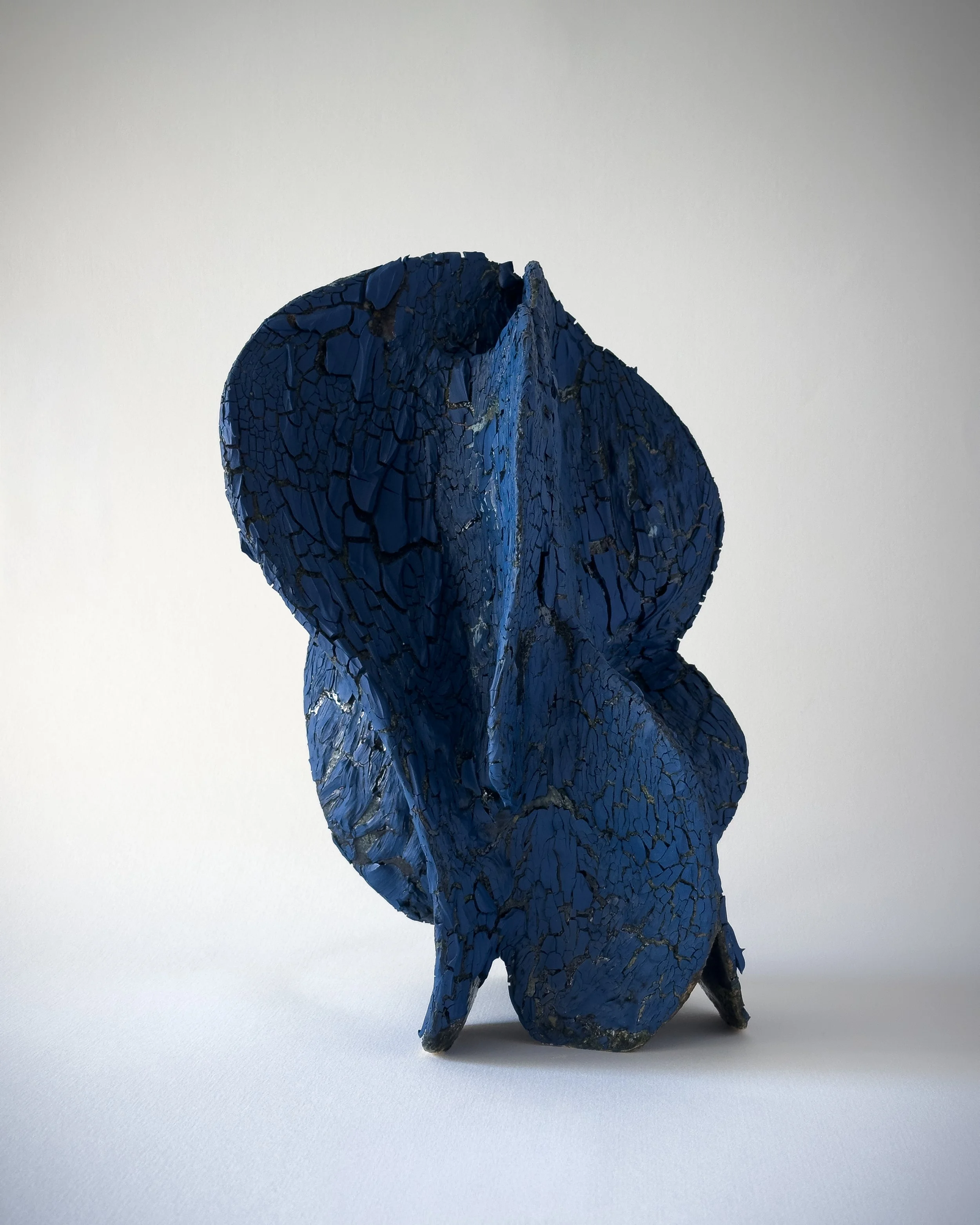 Sculpture made of cracked, painted blue material resembling a stylized face or mask with prominent facial features, standing on a white surface against a plain background.