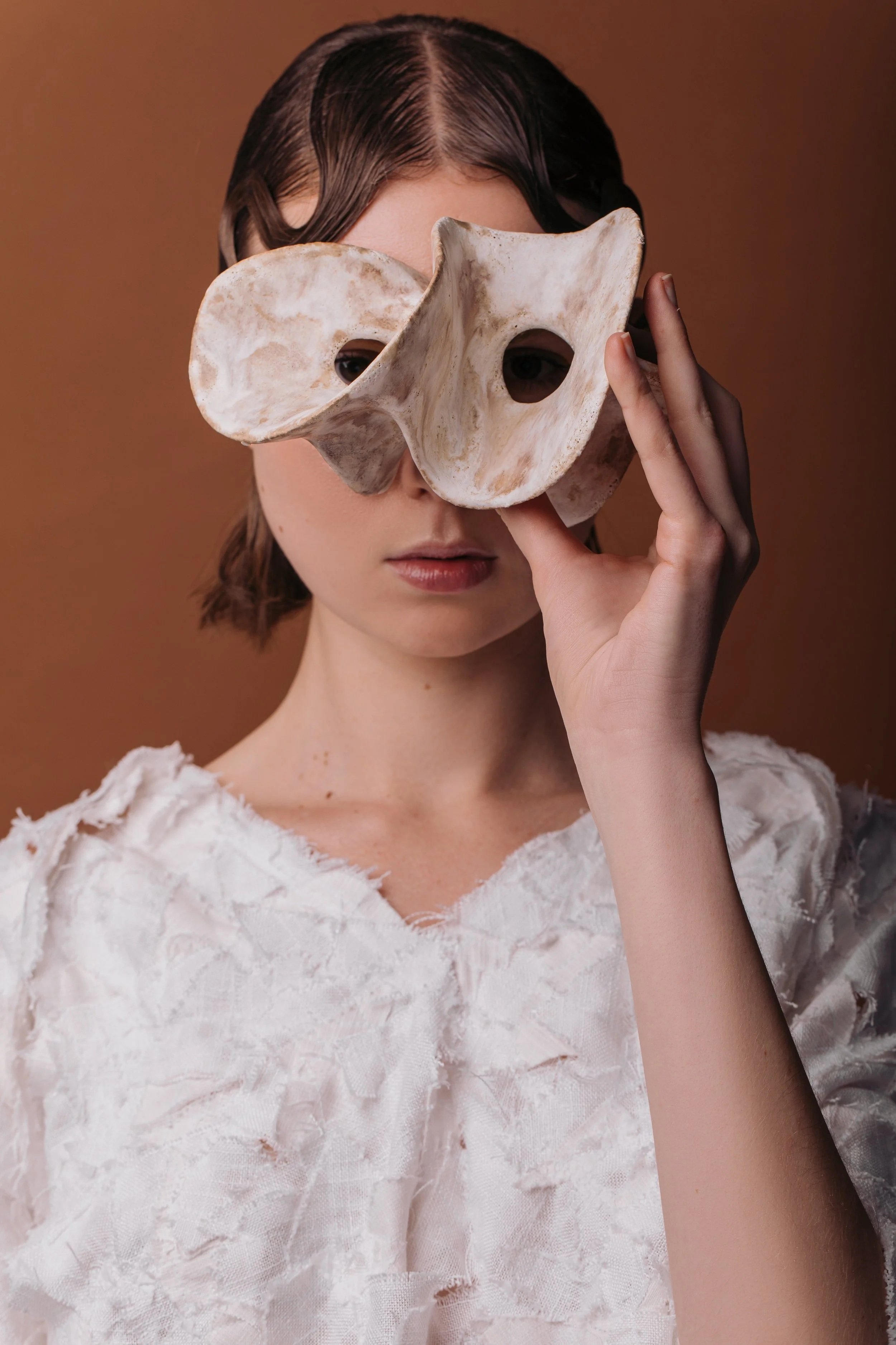 A woman with short brown hair holds a ceramic mask over her eyes, partially covering her face, wearing a white textured top with a brown background.