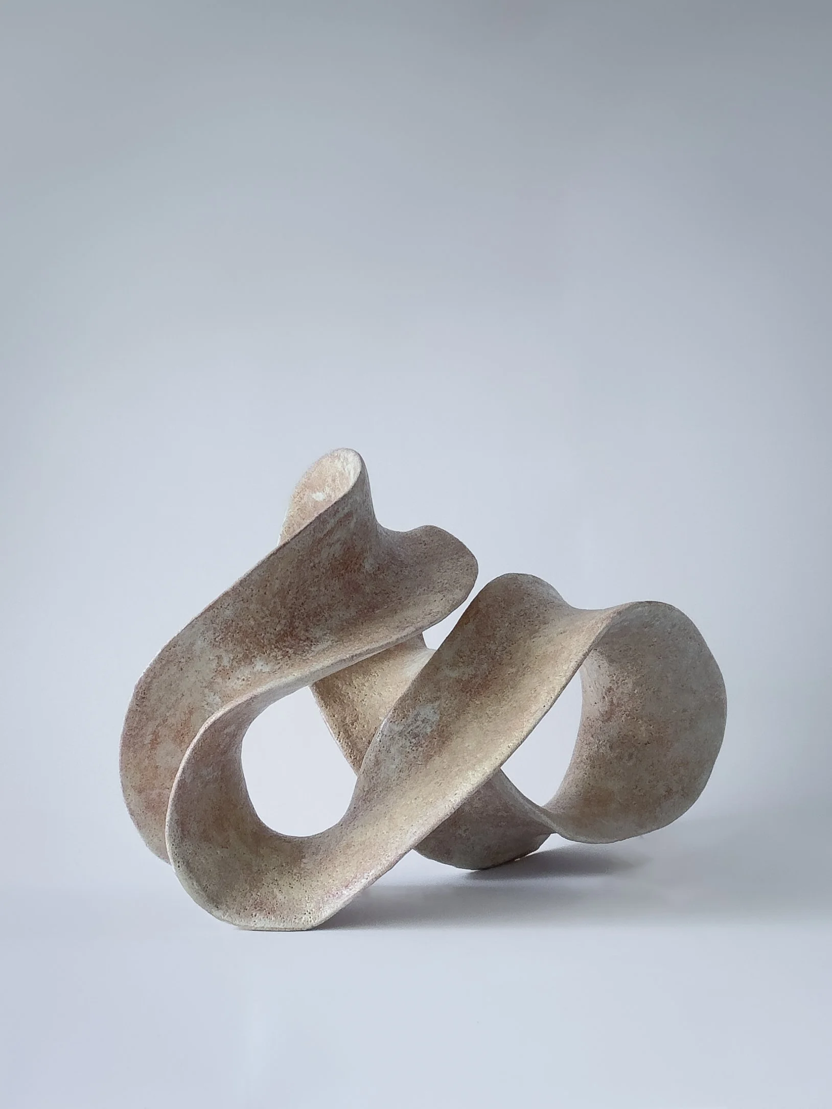 Abstract, curved sculpture made of stone or ceramic with a loop and twisting form, set against a plain light grey background.