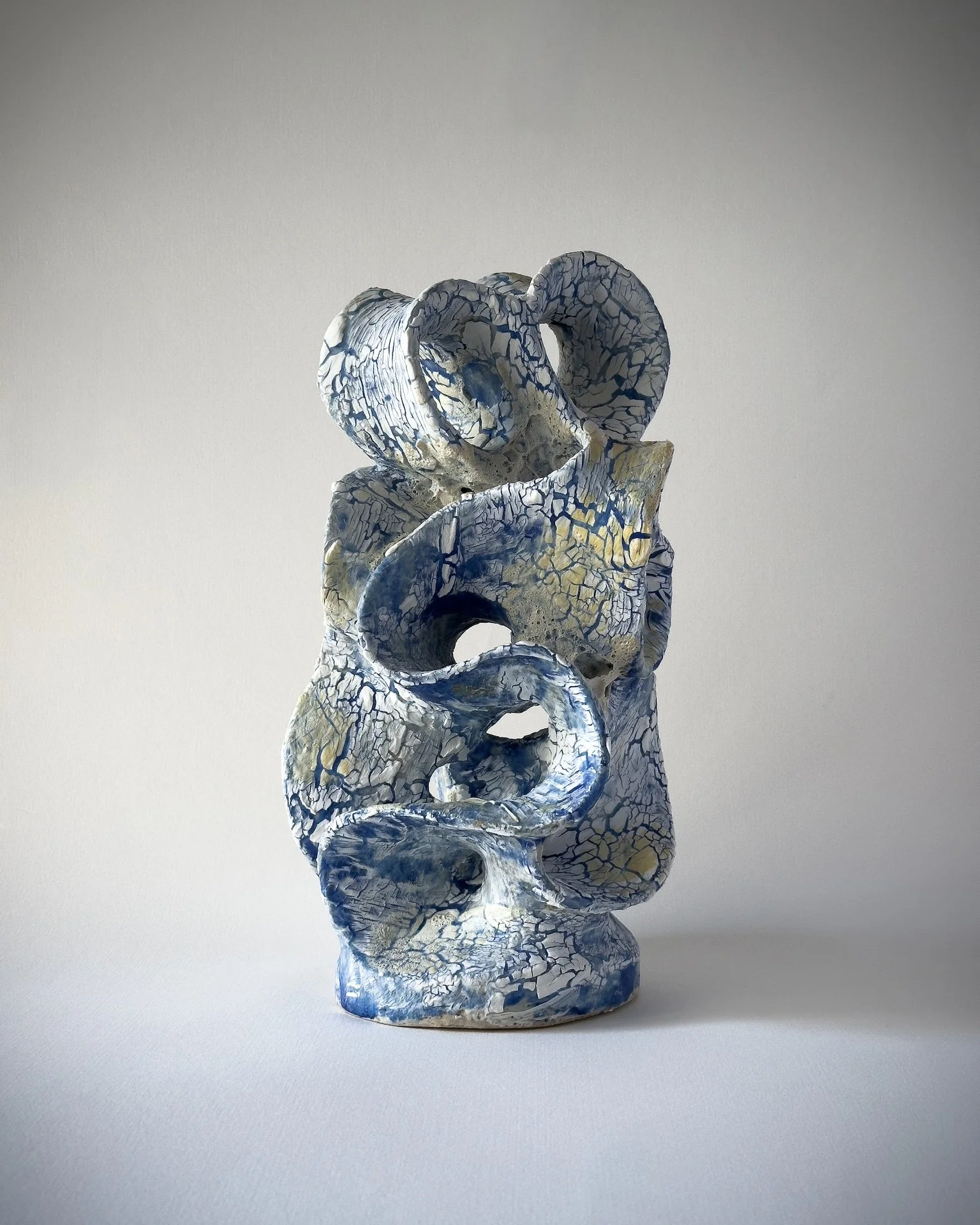 A tall sculpture made of interconnected, cracked ceramic loops with a white, blue, and yellow glaze, set against a plain light background.