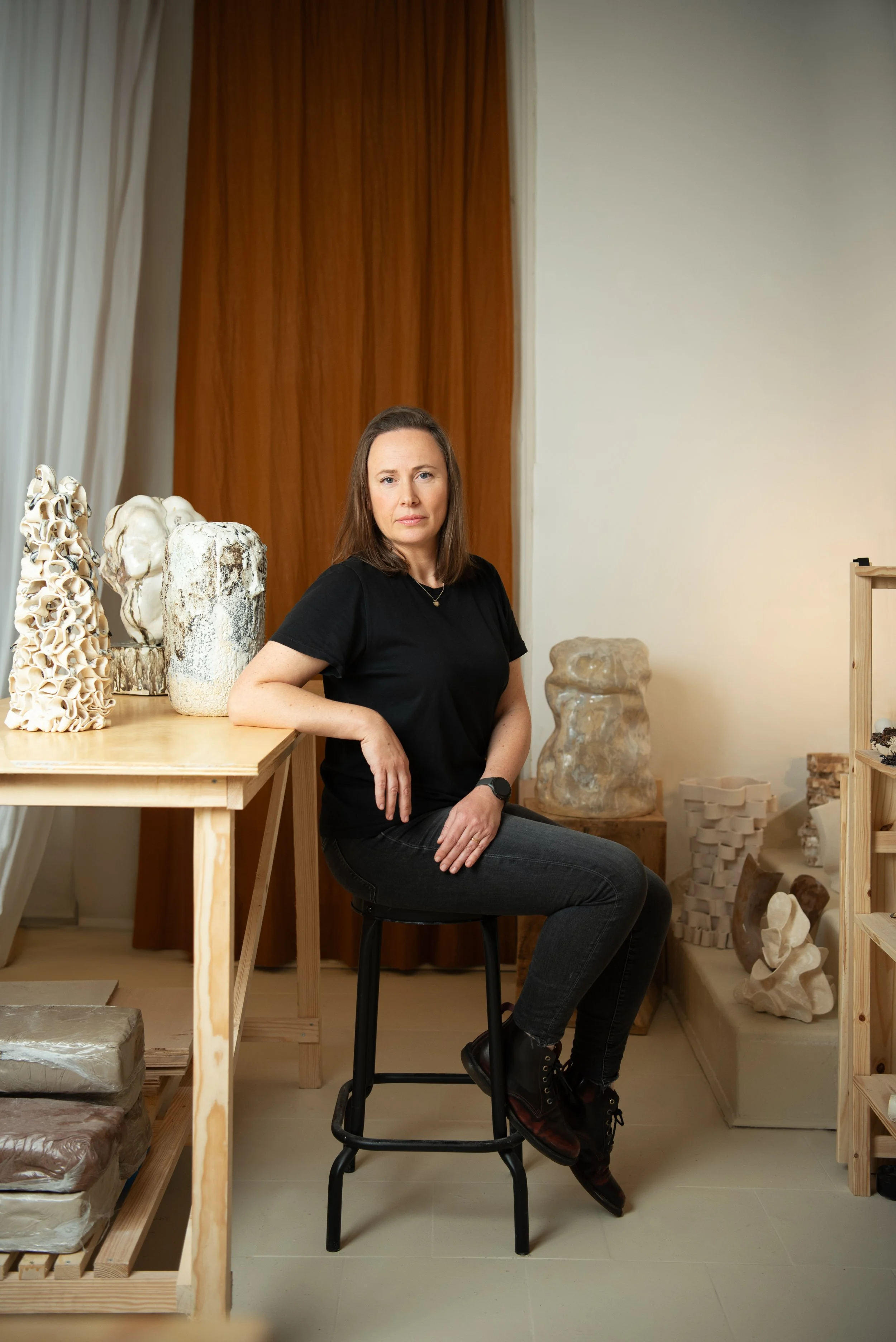 A woman with brown hair wearing a black t-shirt, jeans, and black boots sitting on a black stool in an art studio. She is surrounded by sculptures and pottery on shelves and tables.